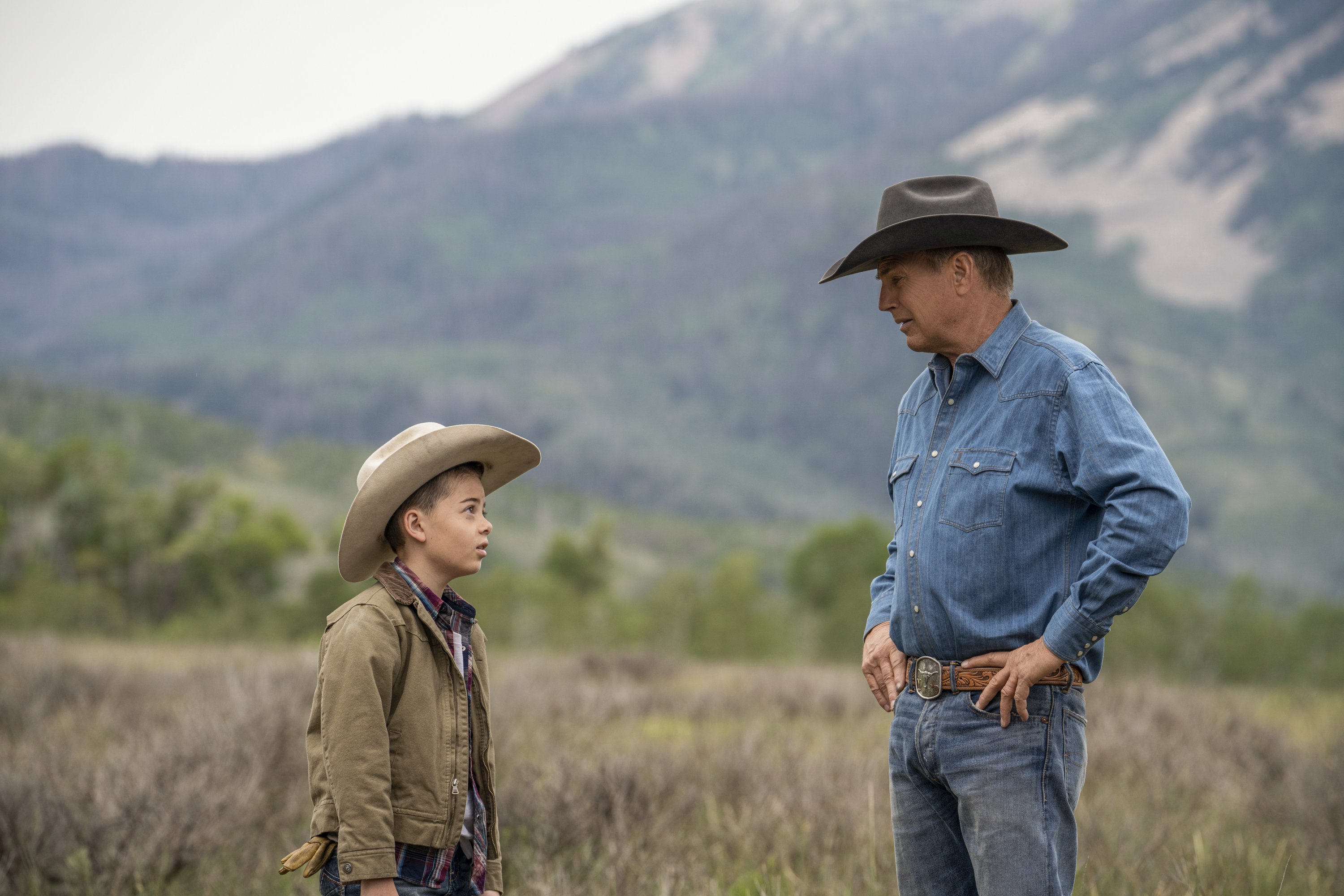 Yellowstone’s John and Tate wear cowboy hats and stand in tall grass, mountains in the background.
