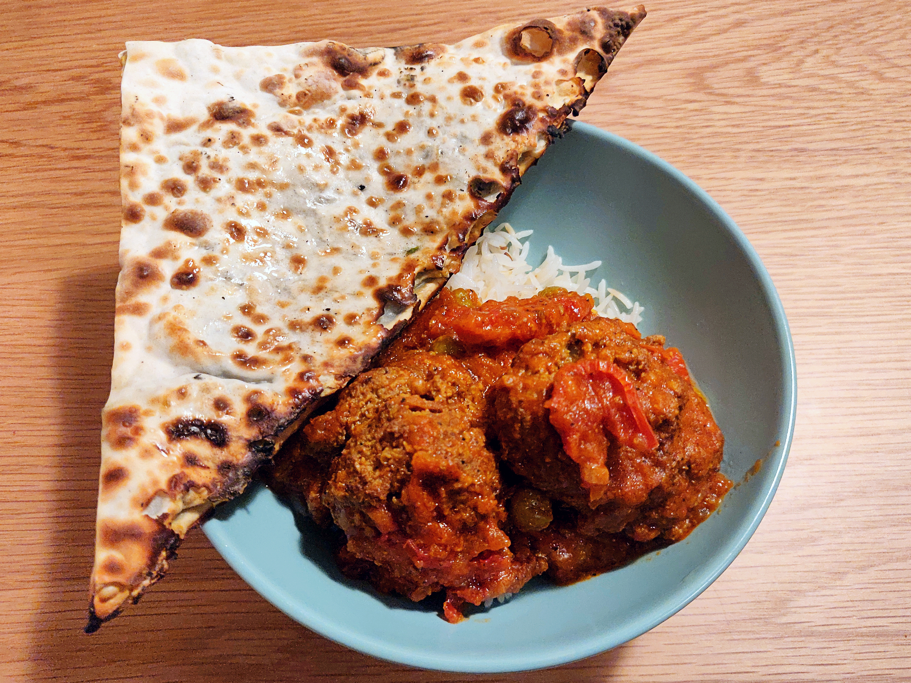 Two plump meatballs in red sauce sit on a bed of white rice, with a thin, crispy triangle of flatbread on the side.