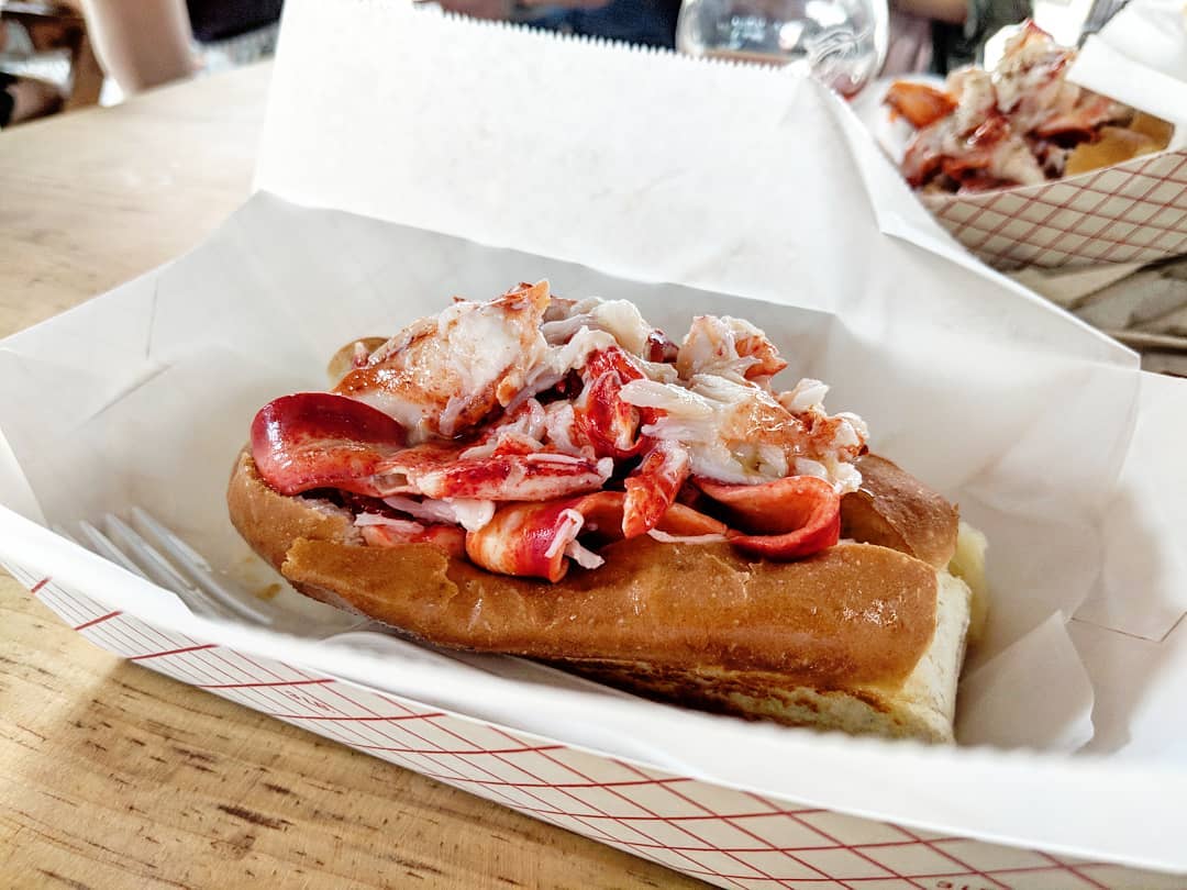 A hearty pile of lobster meat, coated with butter, is stuffed into a New England-style hot dog roll in a red-and-white paper tray.