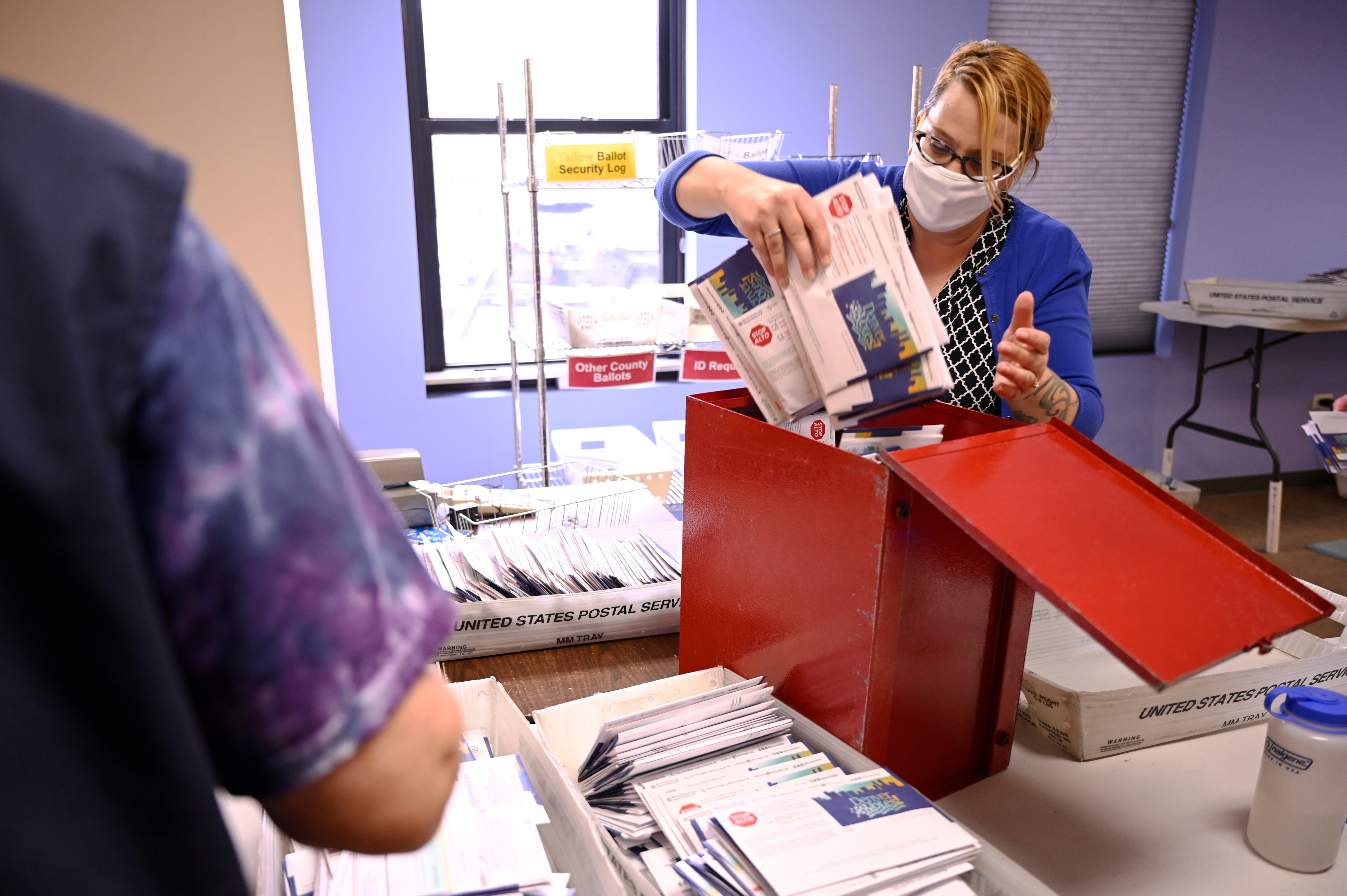 A voting services manager, wearing a mask and blue jacket, pulls a stack of ballots out of a red ballot box, around several bins of sorted ballots.