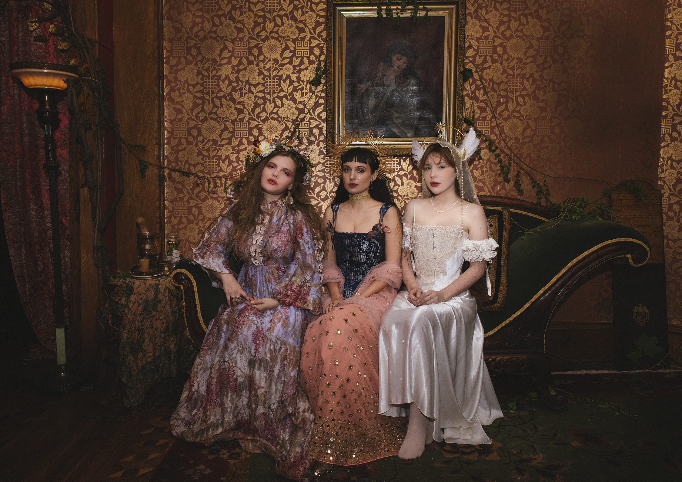 Three young women in romantic dresses sit on an ornate sofa in a richly decorated room.