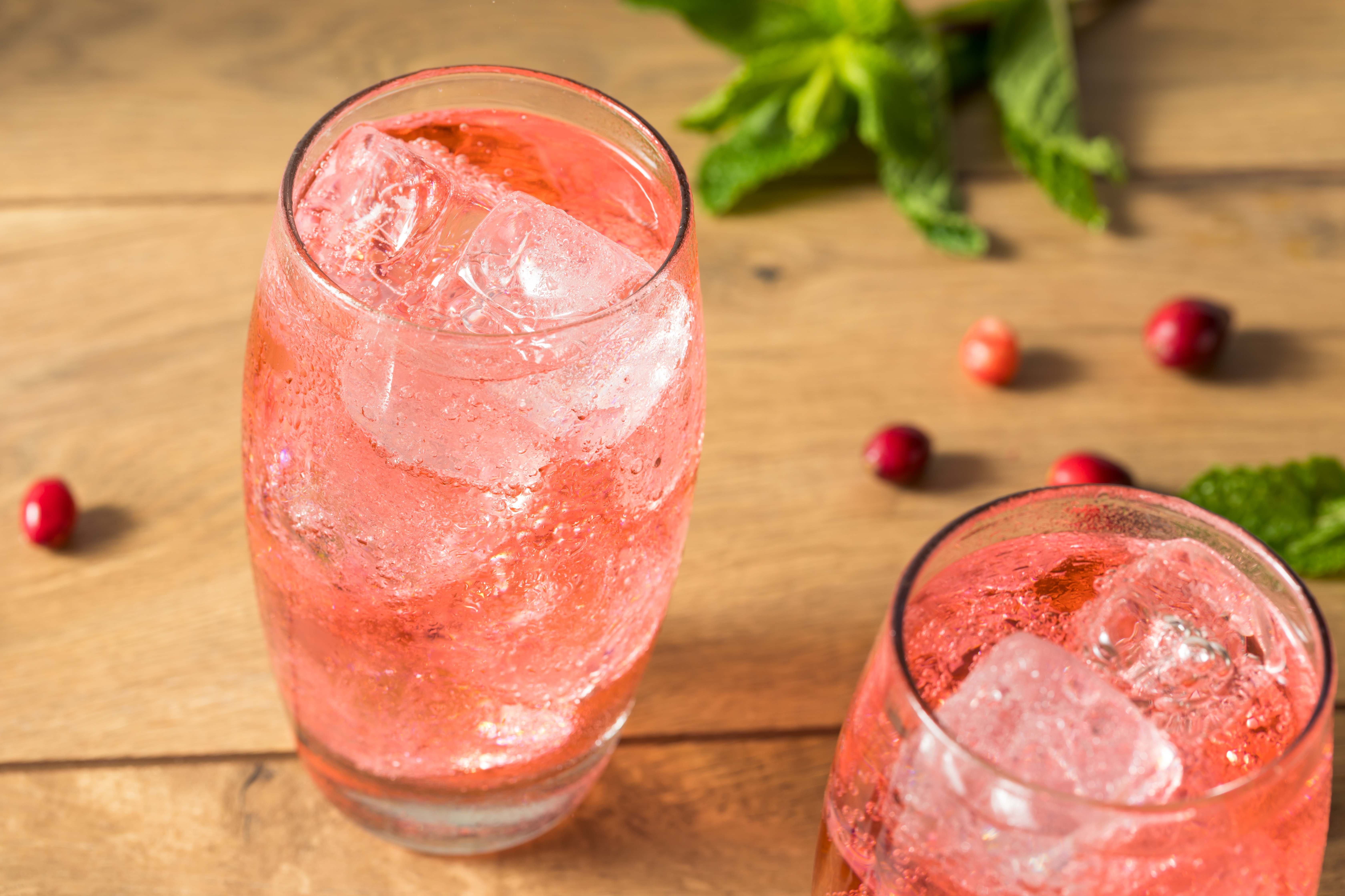 Two curved highball glasses full of ice cubes and a light pink soda are set on a wood table, surrounded by scattered cranberries and leaves.