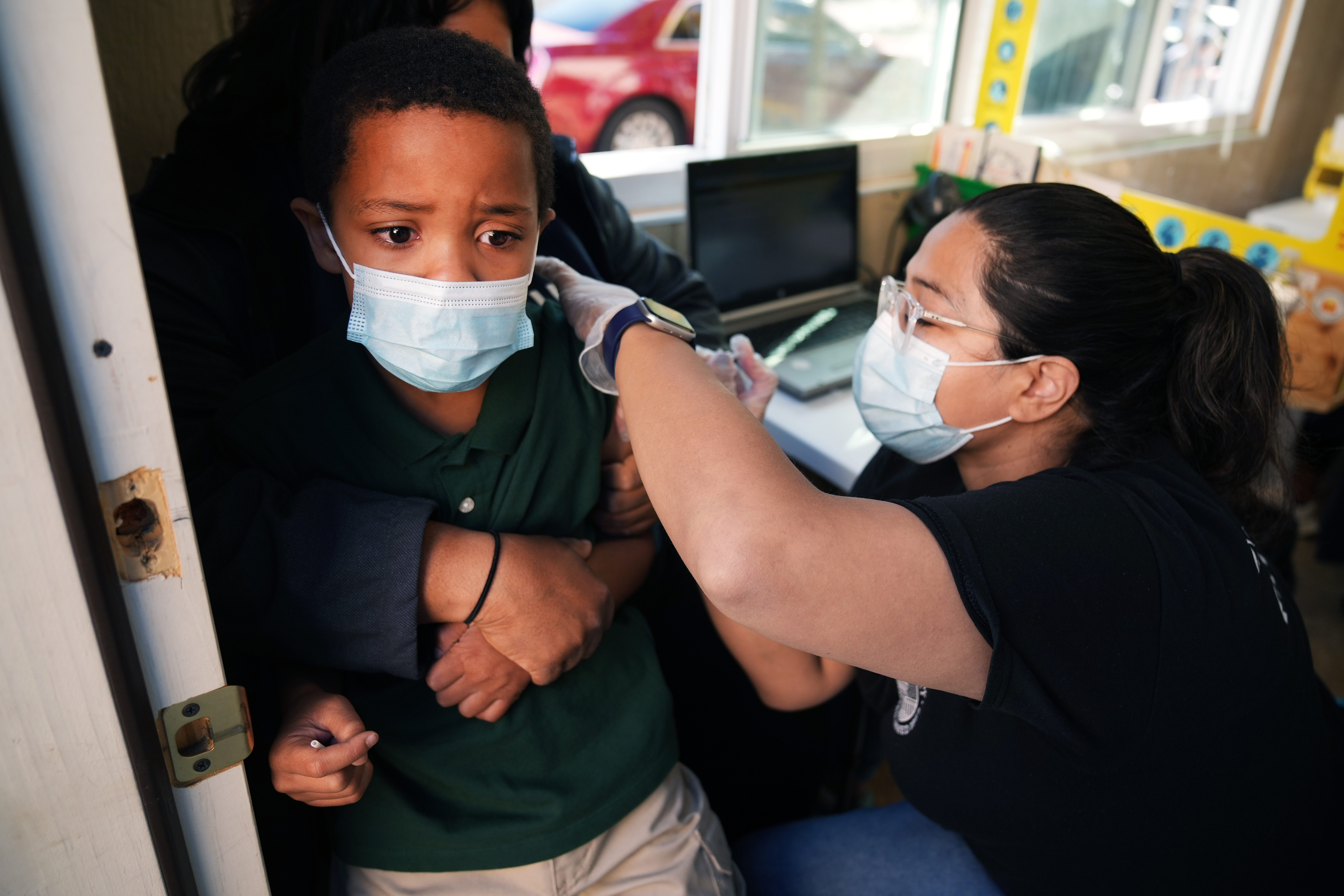 A health care professional wearing glasses and a white protective mask gives a dose of COVID vaccine to a young boy, who is being held by a parent.