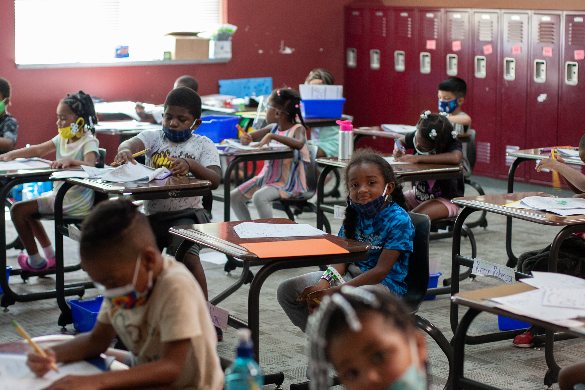 About 10 young kindergarten students sit at small desks looking at worksheets in a classroom lined with red lockers. One girl in a blue shirt and a black face mask in the middle of the photo looks at the camera.
