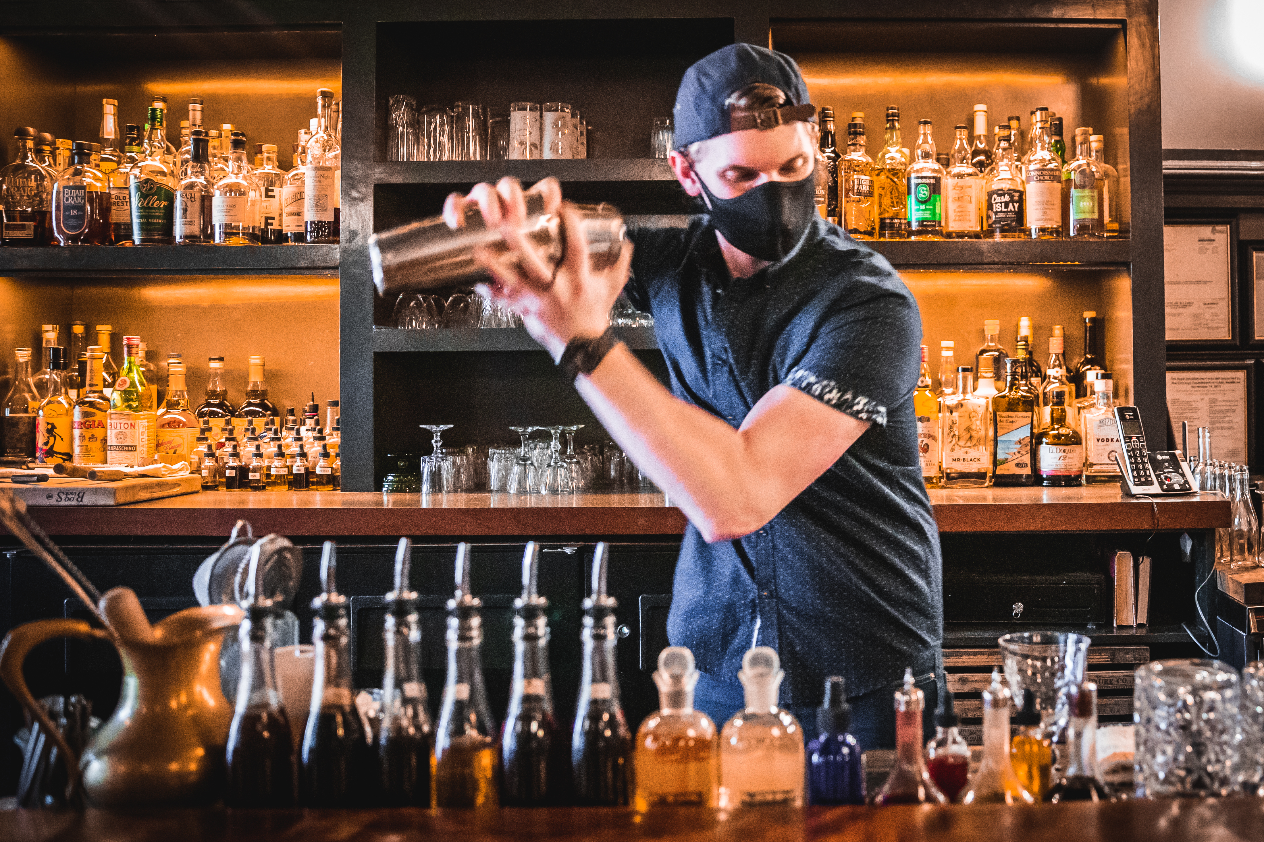 A bartender wearing a black face mask shakes a cocktail behind a bar.