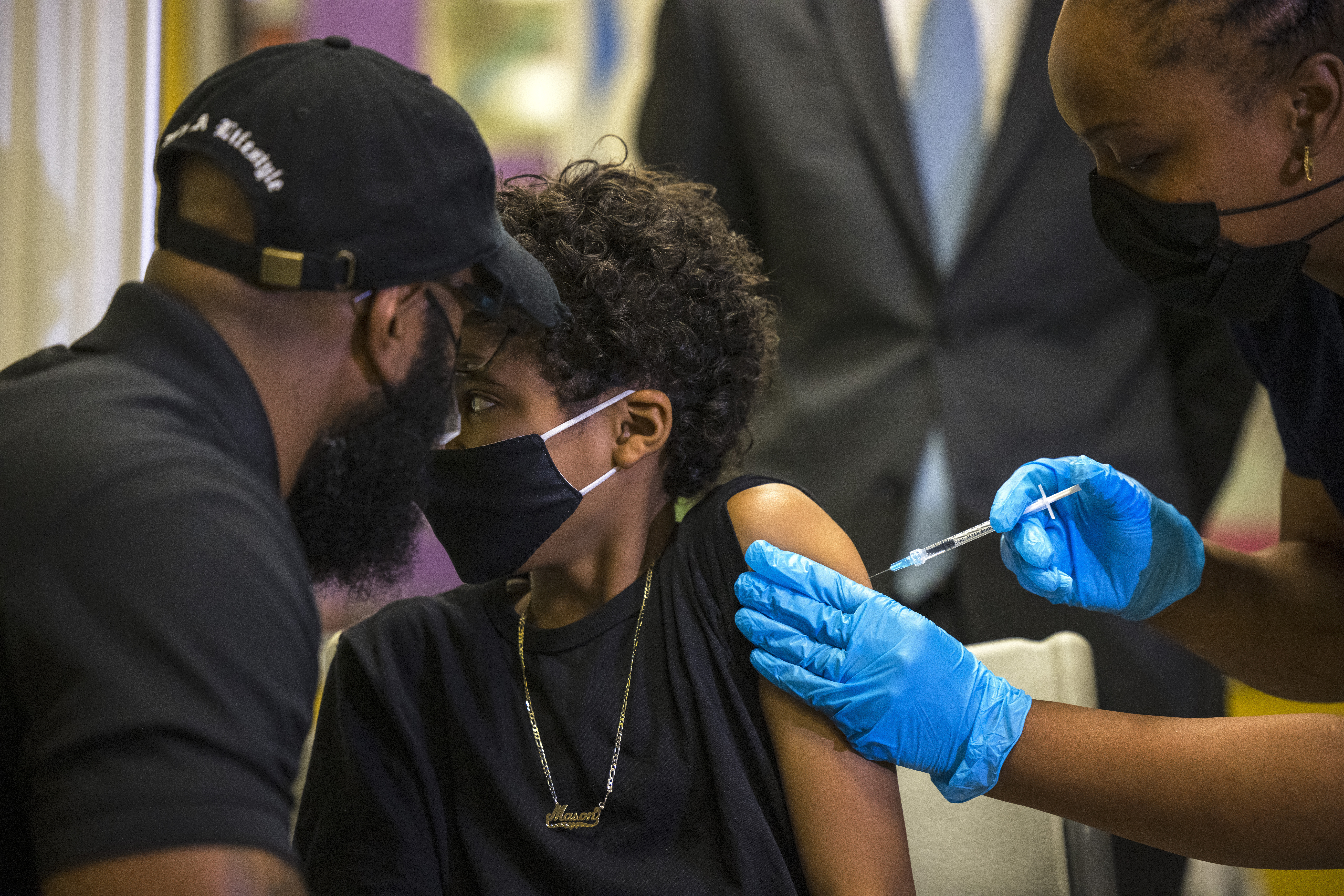 A boy receives a dose of a COVID vaccine from a medical professional wearing bright blue latex gloves. A man wearing a black shirt and hat sits in front of the child.