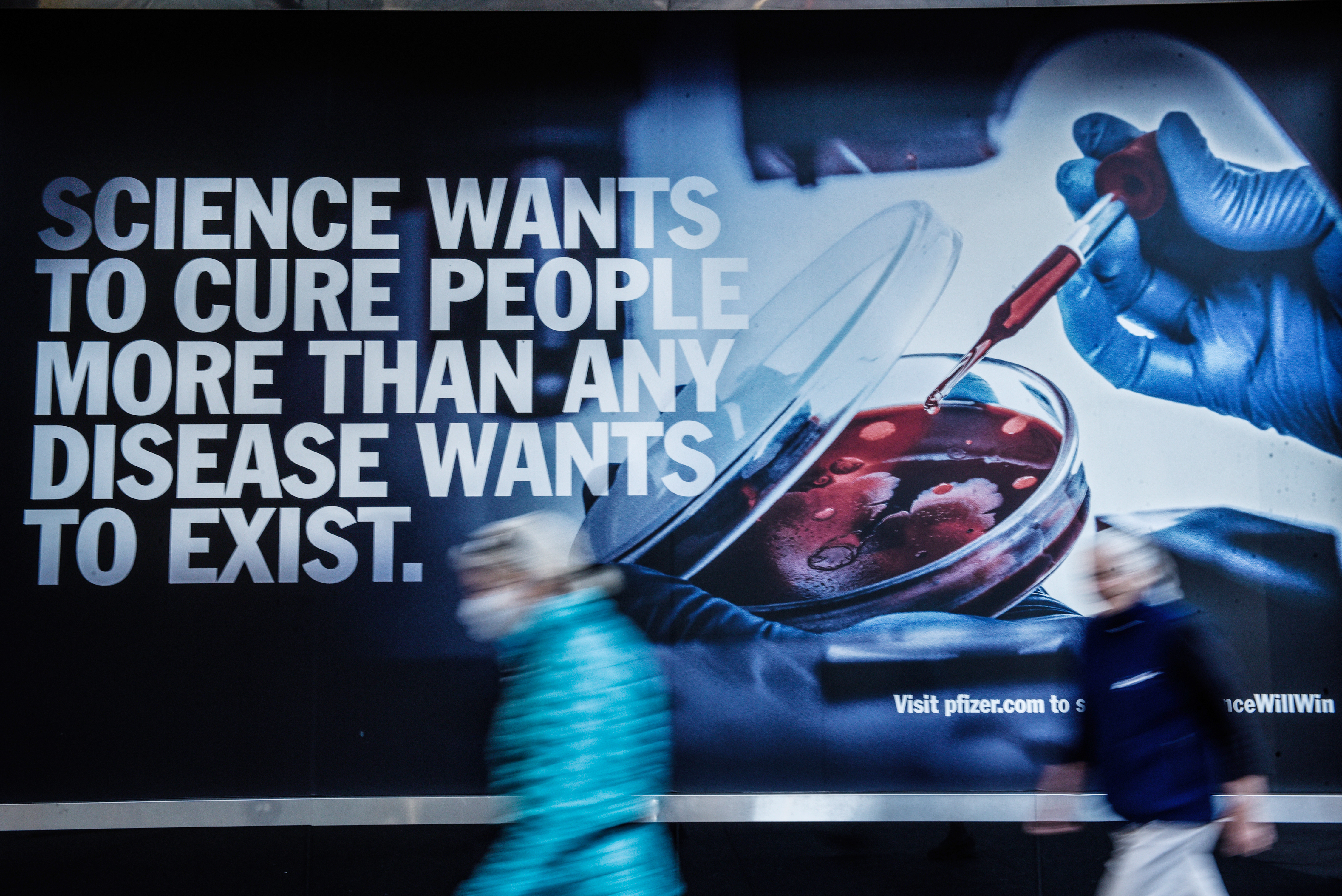 Pedestrians walk past a wall billboard that reads “Science wants to cure people more than any disease wants to exist.”