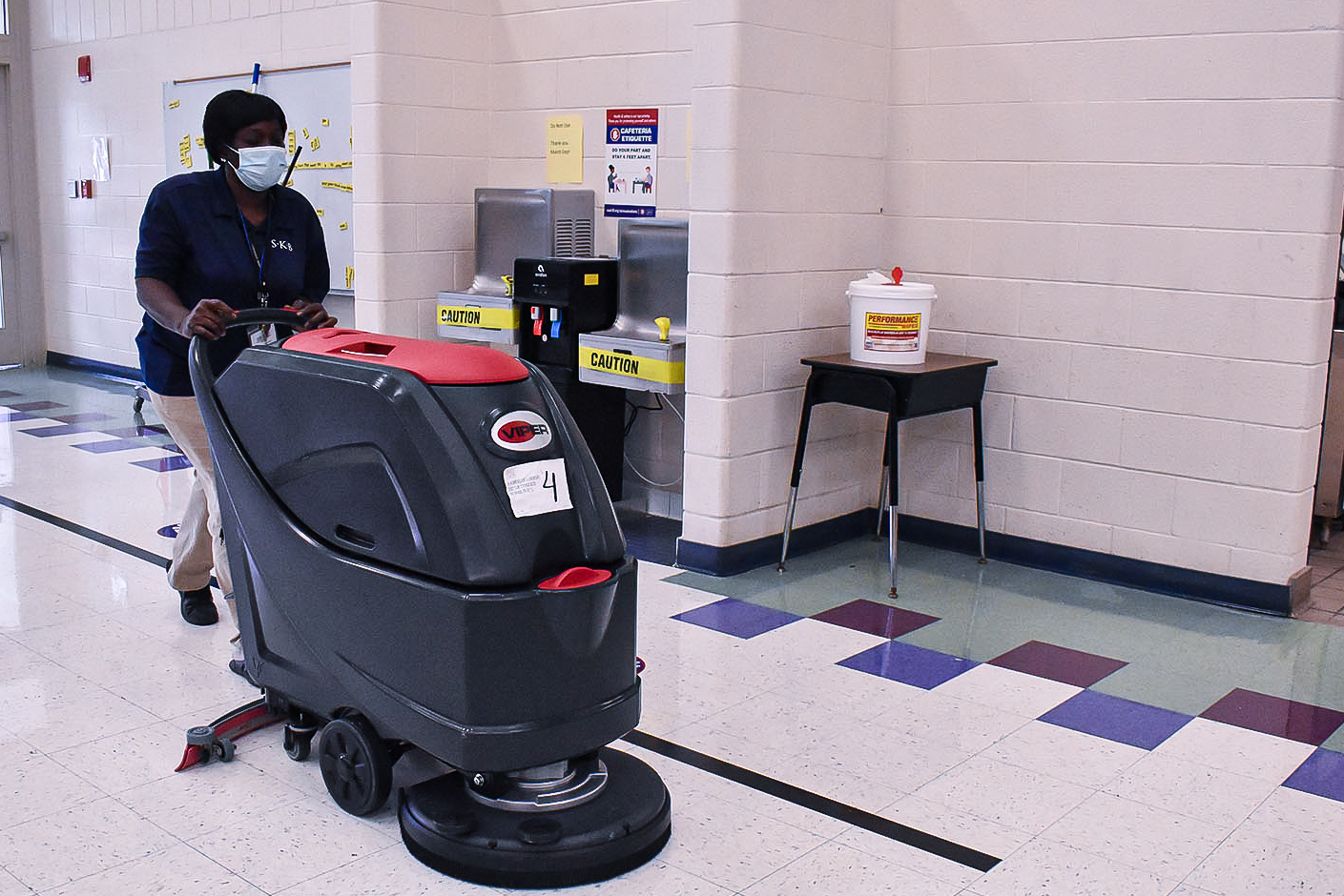 A Shelby County Schools employee, wearing a blue polo and blue protective mask, pushes a floor buffer down a tiled hallway.