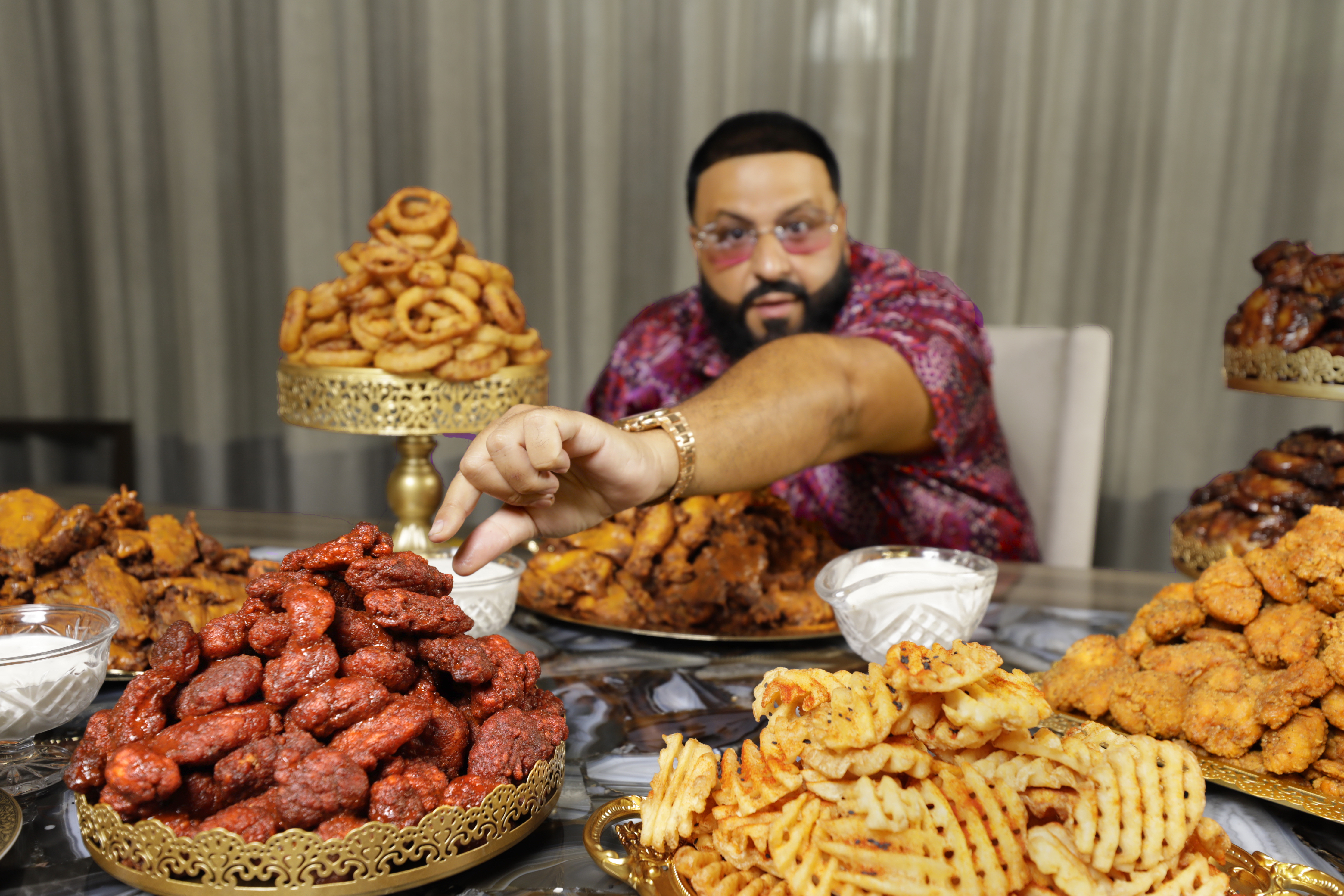 A man wearing sunglasses sitting at a table full of chicken wings, onion rings, and fries.