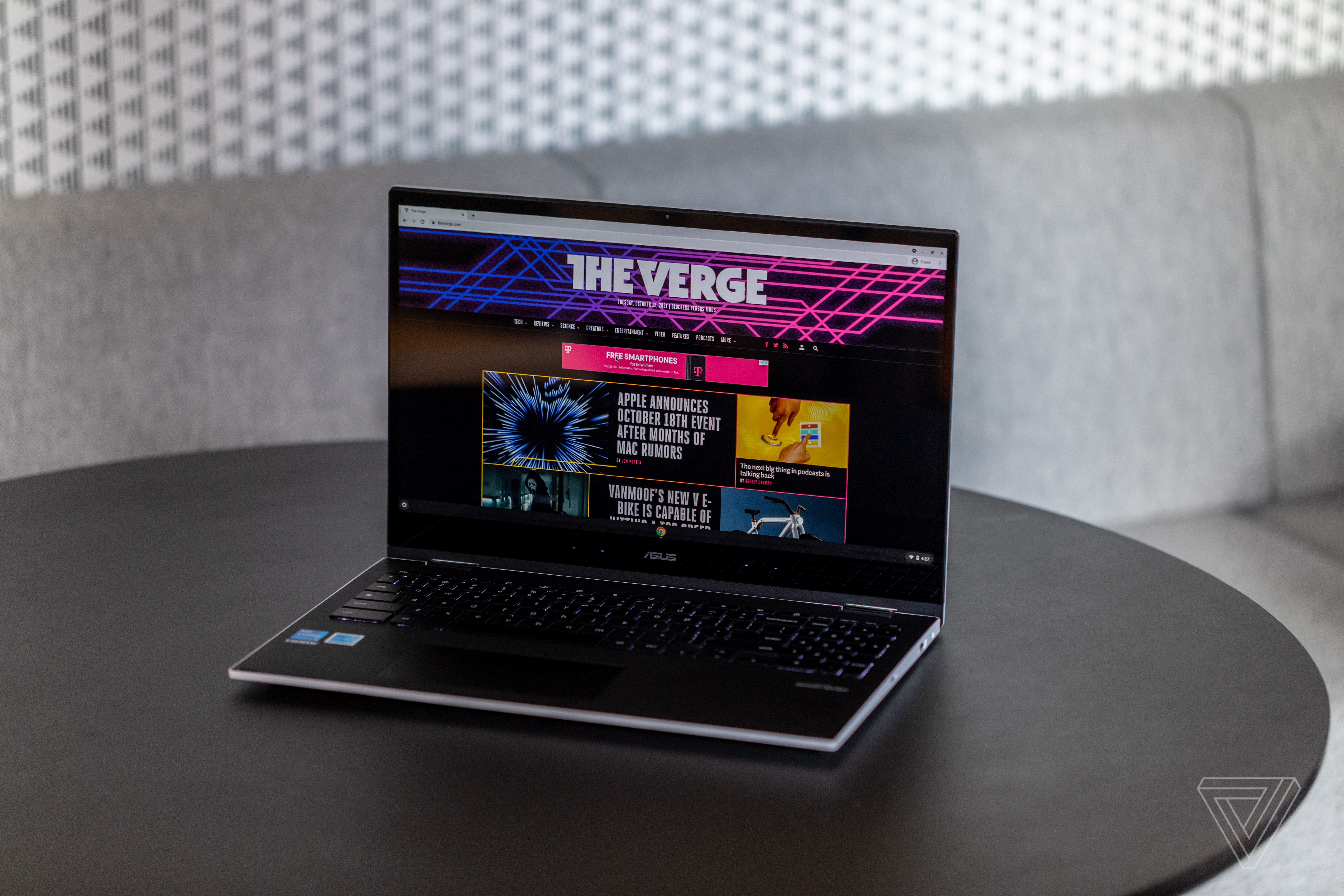 The Asus Chromebook Flip CX5 open on a black table, angled to the left, with a gray fabric bench and white textured wall in the background. The screen displays The Verge homepage.