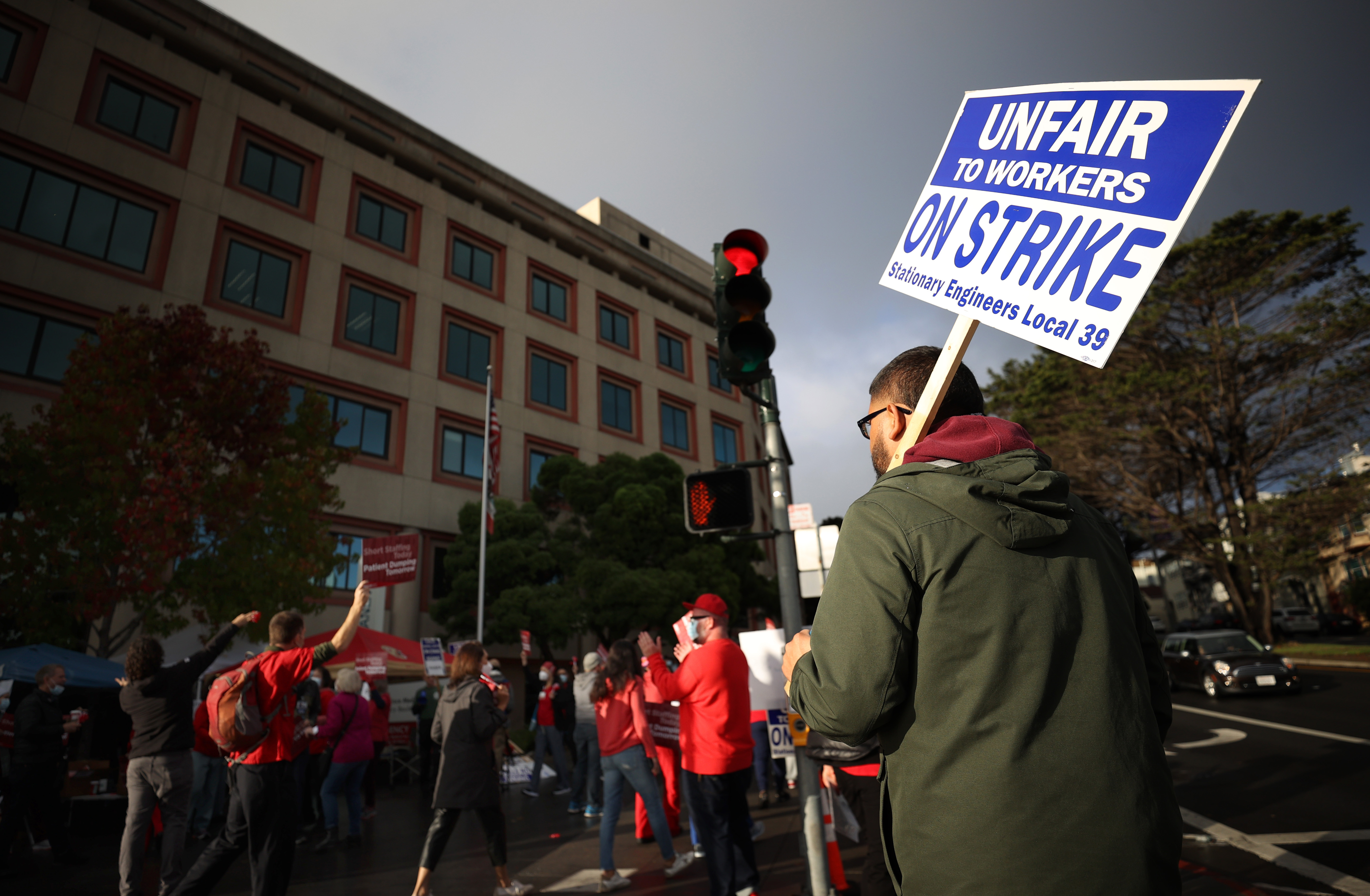 A picketer on a city street carries a sign that reads “Unfair to workers, on strike.”