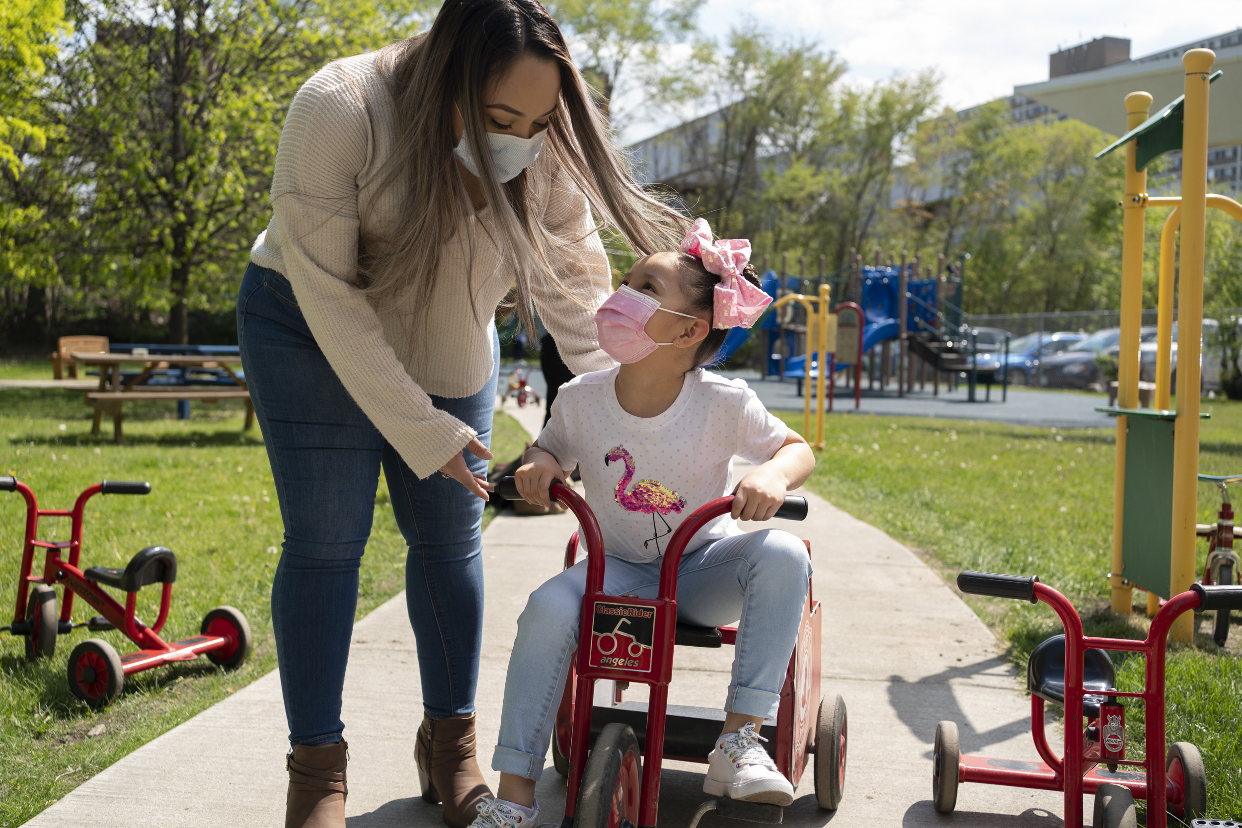 A mother wearing jeans, tan shirt and protective mask pushes her daughter on a red tricycle. The daughter is wearing a white flamingo shirt, pink mask, pink bow and jeans.