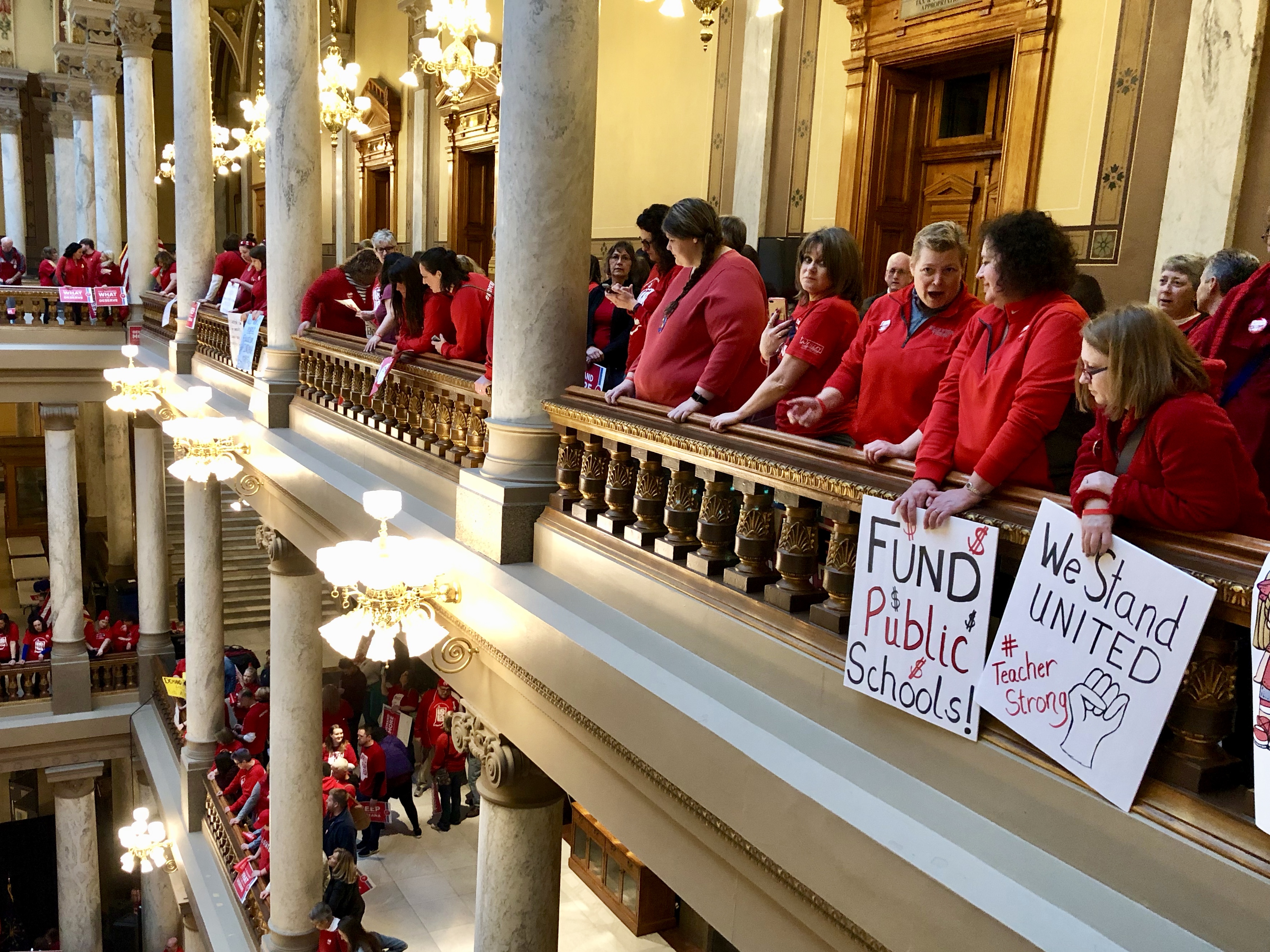 A row of teachers wearing red stand in the balcony of the state Capitol on March 9, 2019, at a rally hosted by the Indiana State Teachers Association. Two hold placards saying “Fund Public Schools” and “We Stand United.”