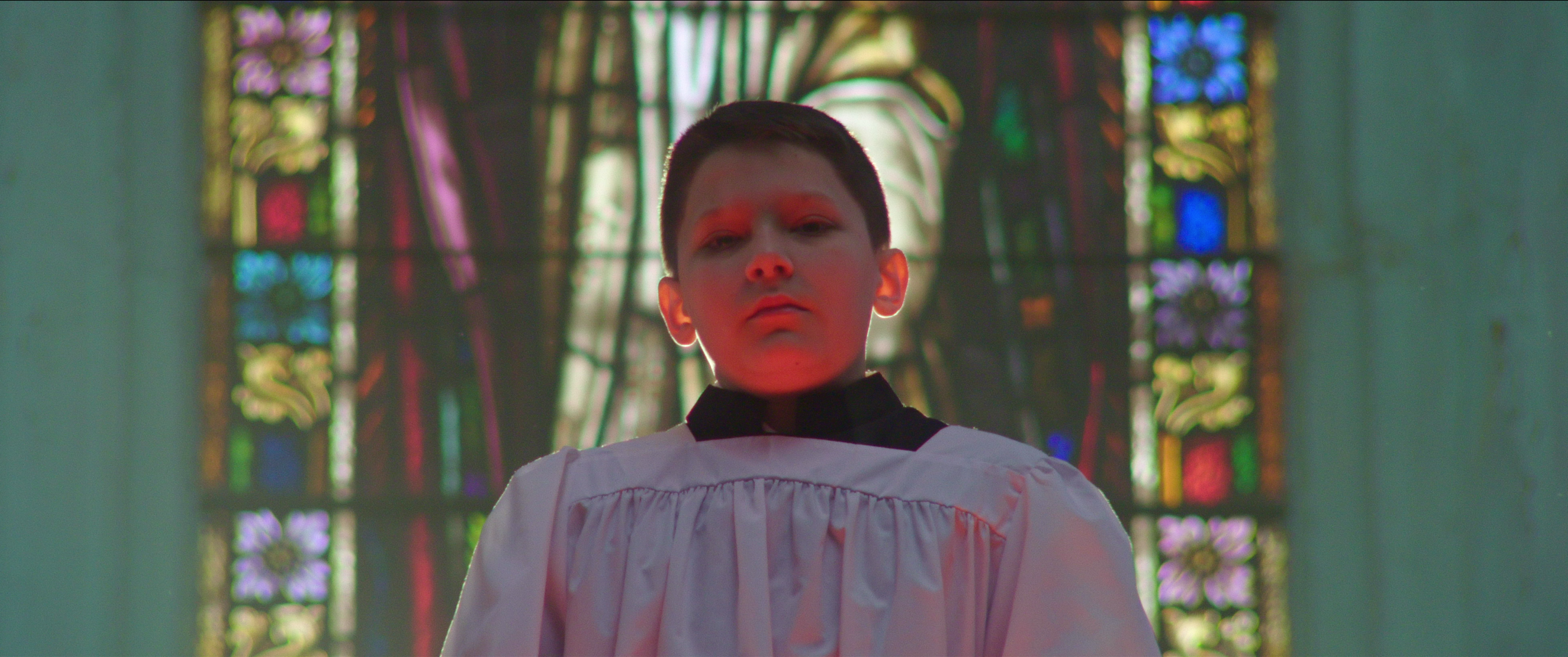 A boy in an altar boy’s robe stands backlit by a stained glass window.
