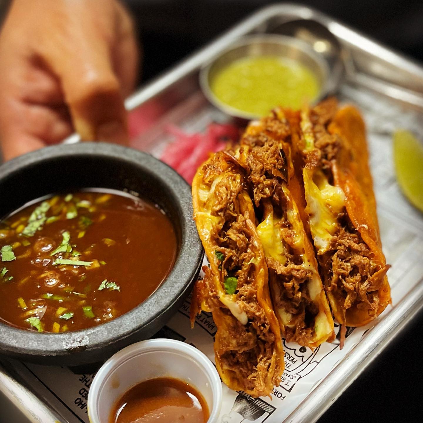 a hand holding a metal, newspaper-lined tray of three yellow-cheese-laced, chicken quesabirria crispy tacos with a side of deep brown, cilantro and scallion-flecked sauce.