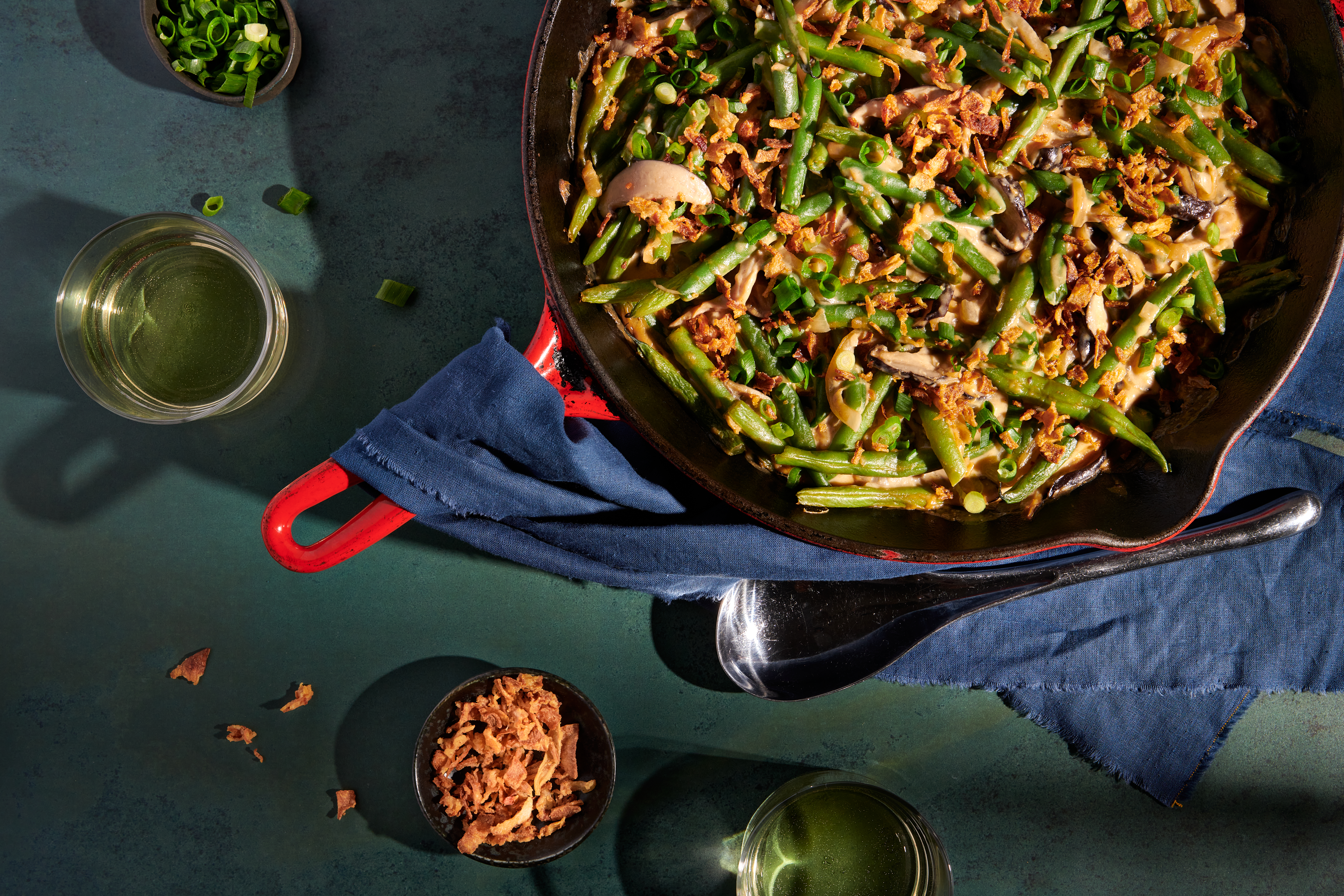 Green bean casserole in a red castiron skillet. Next to the skillet are two drinking glasses, small bowls of sliced scallions and fried shallots. A blue napkin is draped over the skillet handle.