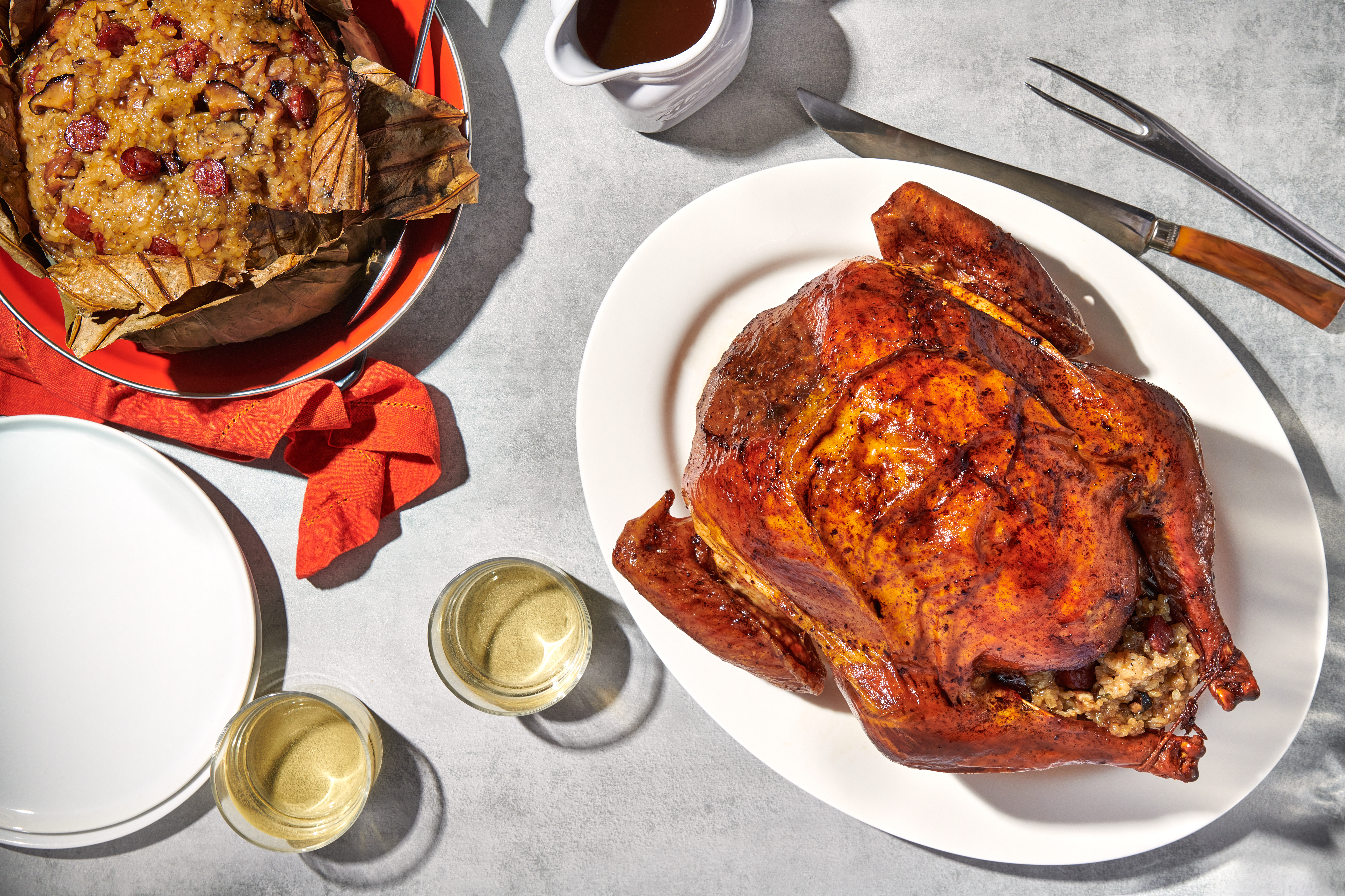 A roasted turkey sits on a white platter next to a big bowl full of sticky rice stuffing. Next to them are two glasses of white wine and serving utensils.