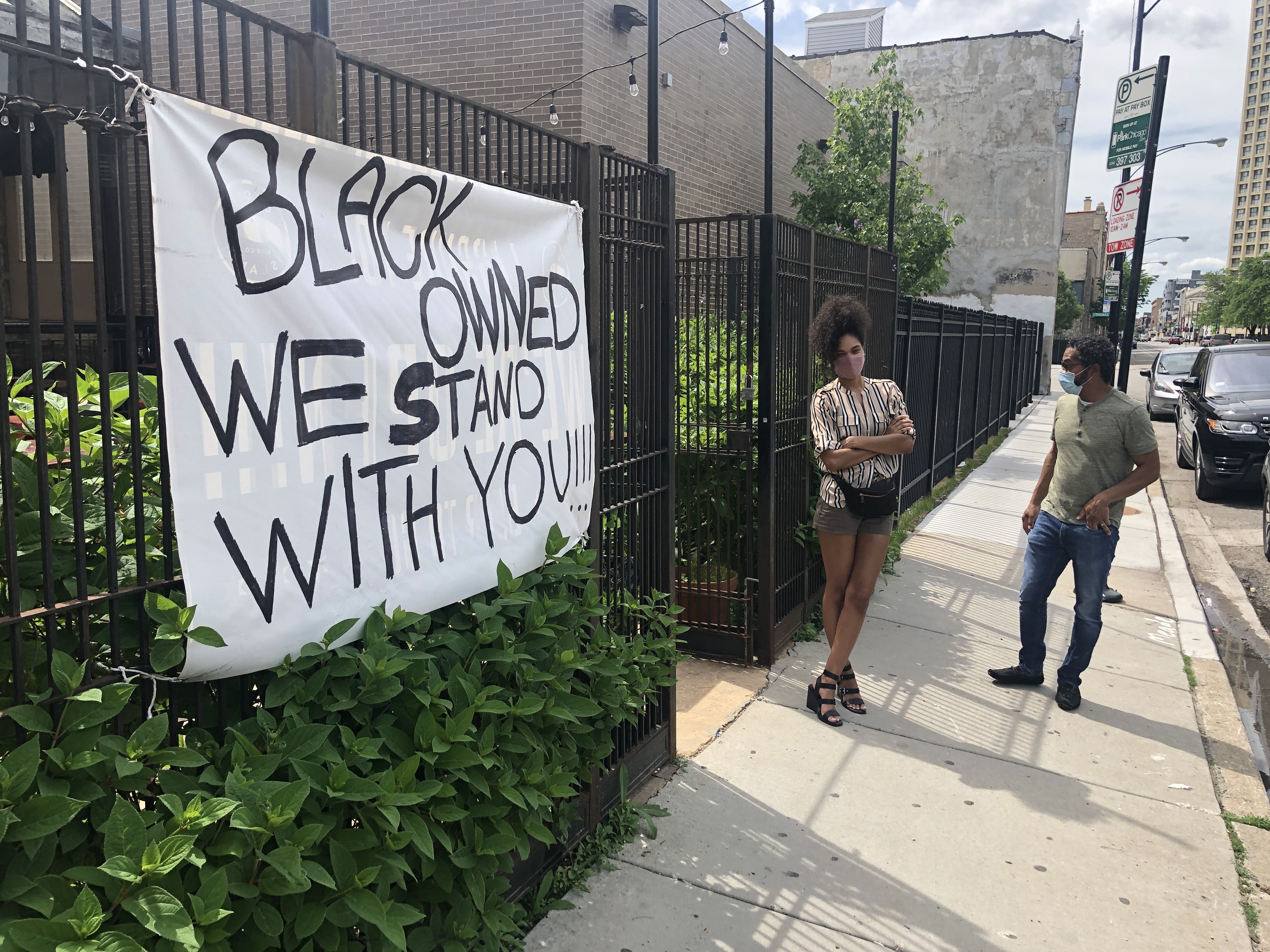 Two people stand outside a black fence where a sign hangs. It reads “Black owned, we stand with you.”