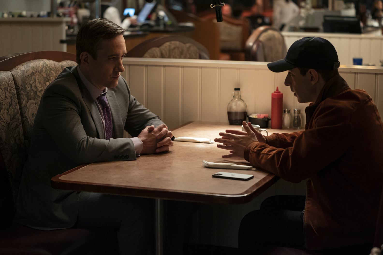 Two men, one in a gray suit and the other in a brown coat and baseball hat, face each other in an upholstered diner booth.