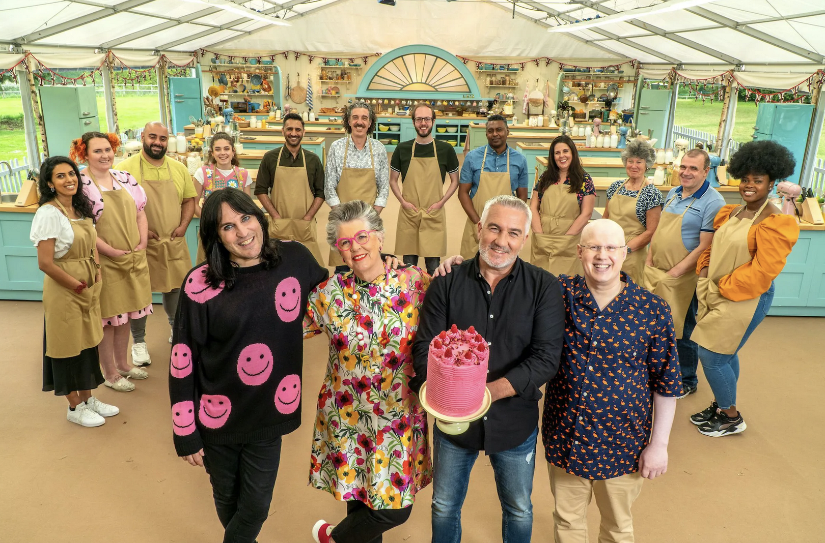 A group portrait of the Great British Bake-Off season 12 contestants, with judges Paul Hollywood and Prue Leith in front alongside hosts Matt Lucas and Noel Fielding. Hollywood holds a big pink layer cake.