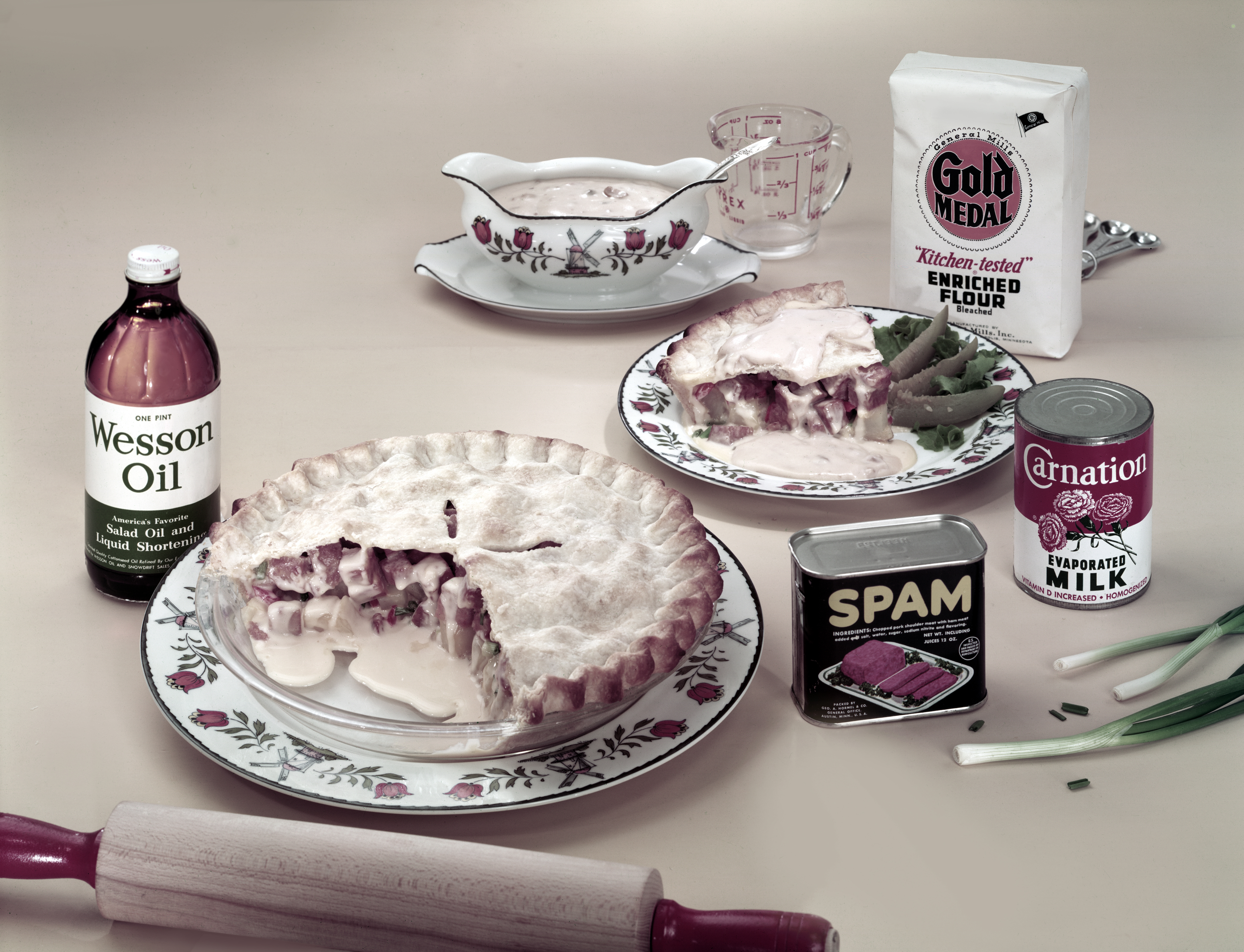 一张50年代或60年代的棕褐色照片，上面是馅饼旁边放着一罐长方形的午餐肉罐头、一罐康乃馨牛奶和威森油。