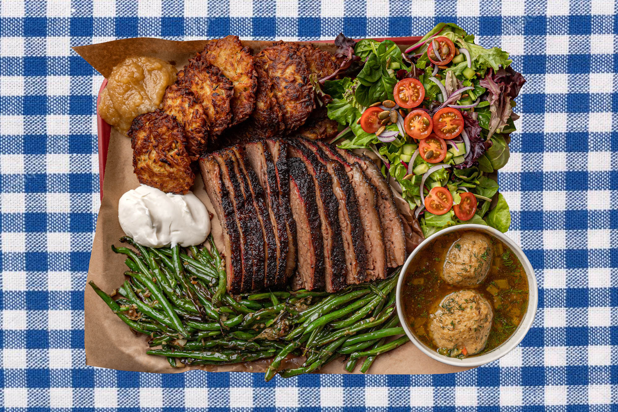 A blue-and-white gingham tablecloth placed underneath a heaping barbecue tray: on top of the tray, thickly sliced brisket, golden-fried latkes, sour cream, green salad, smothered green beans, and matzoh ball soup.