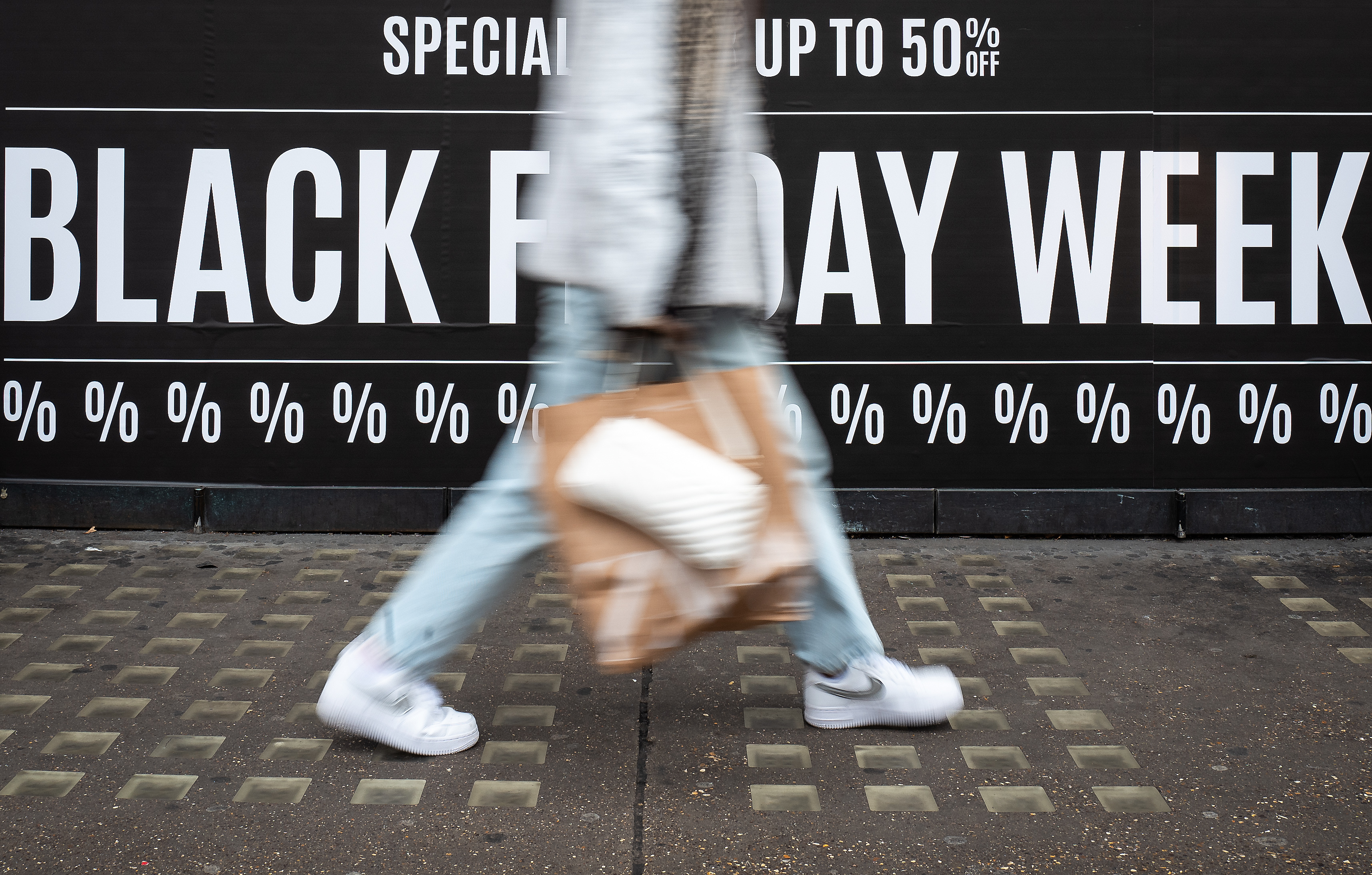 Image of a blurry shopper in London holding a brown bag while walking past an advertisement that says “Black Friday Week.”