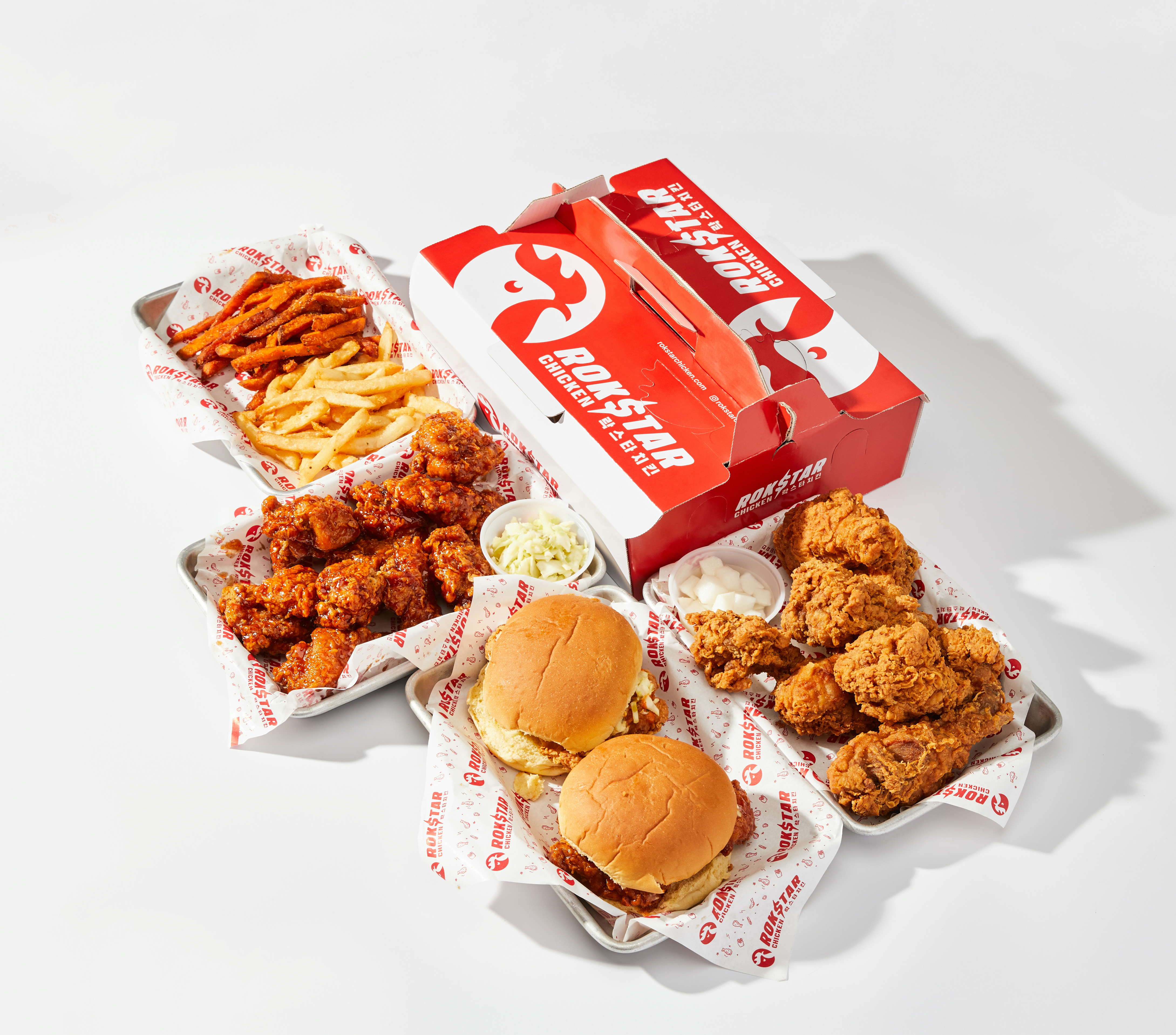 A red and white takeout box and several serving trays filled with fried chicken, two fried chicken sandwiches, and fries.