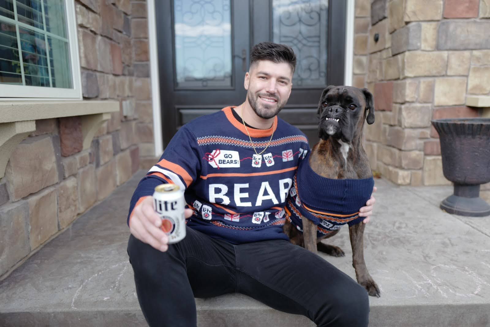 A man sits on a stoop wearing an ugly orange and blue sweater, hugging a dog with an underbite that also wears a blue and orange sweater and holding a can of beer