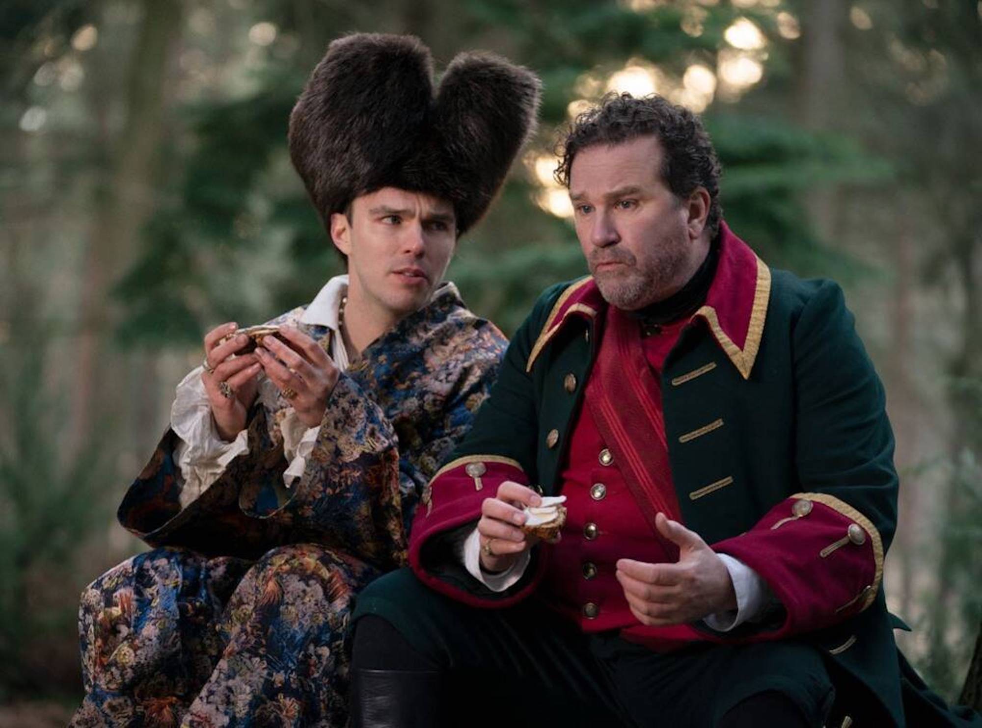 The characters Peter (played by Nicholas Hoult), dressed in a tall fur hat and brocaded coat, and General Velementov (Douglas Hodge), in an 18th century military uniform, sit in the woods eating cheese on bread.