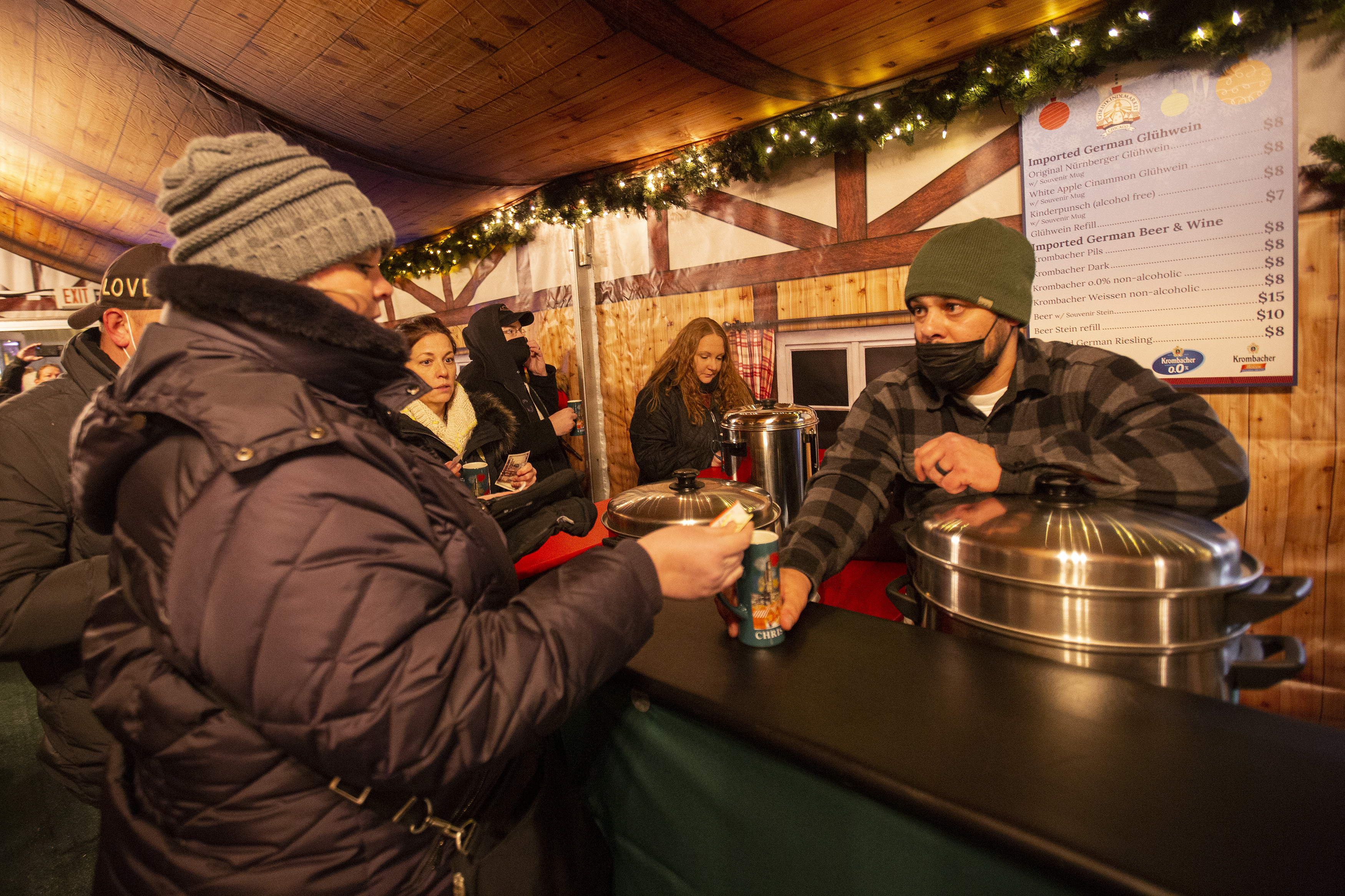 A man hands a mug of hot, mulled wine to a woman from behind a wooden outdoor bar.