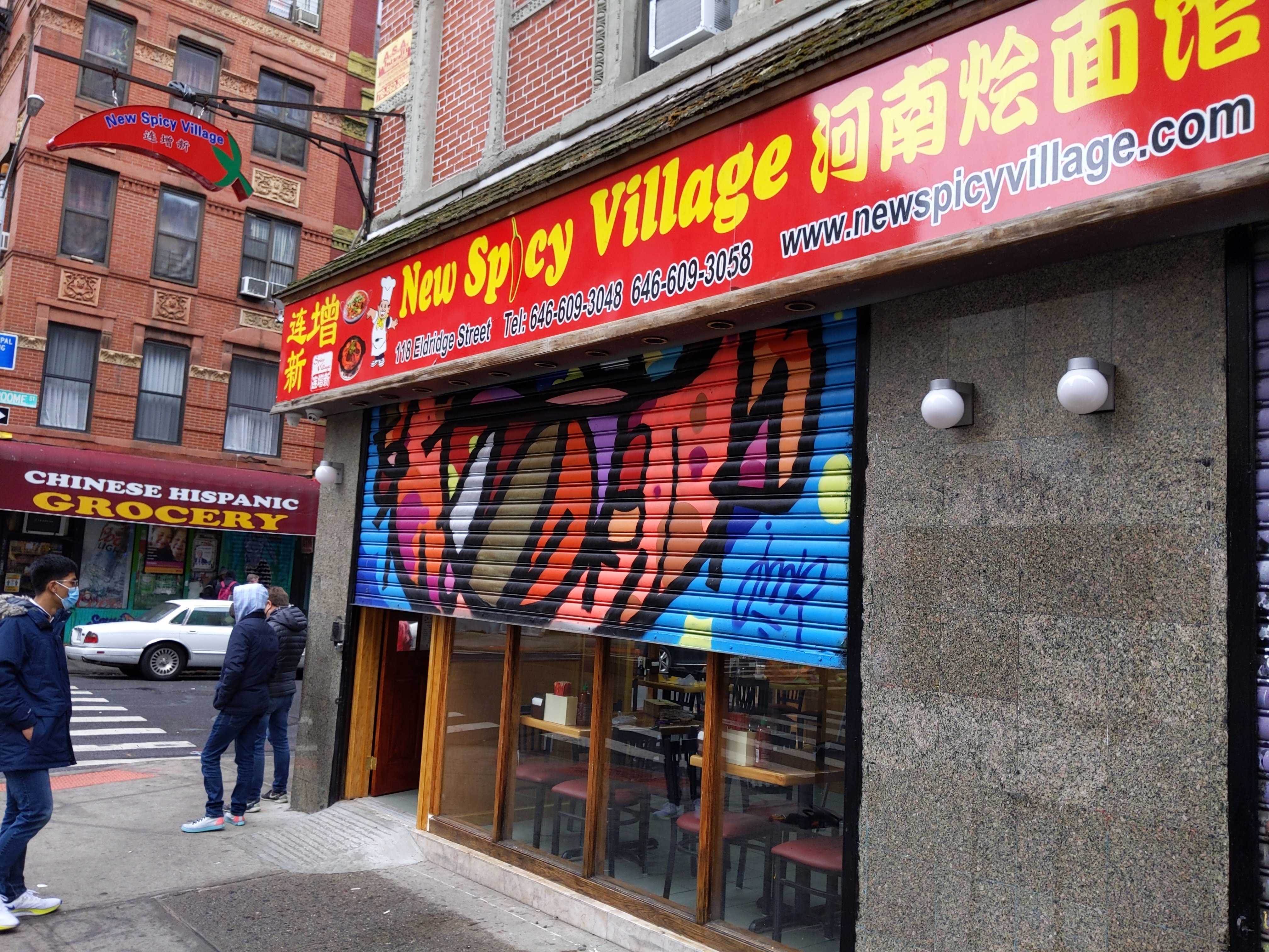 A red exterior restaurant sign with yellow lettering spelling out New Spicy Village. A graffitied gate is halfway covering the restaurant’s front windows.