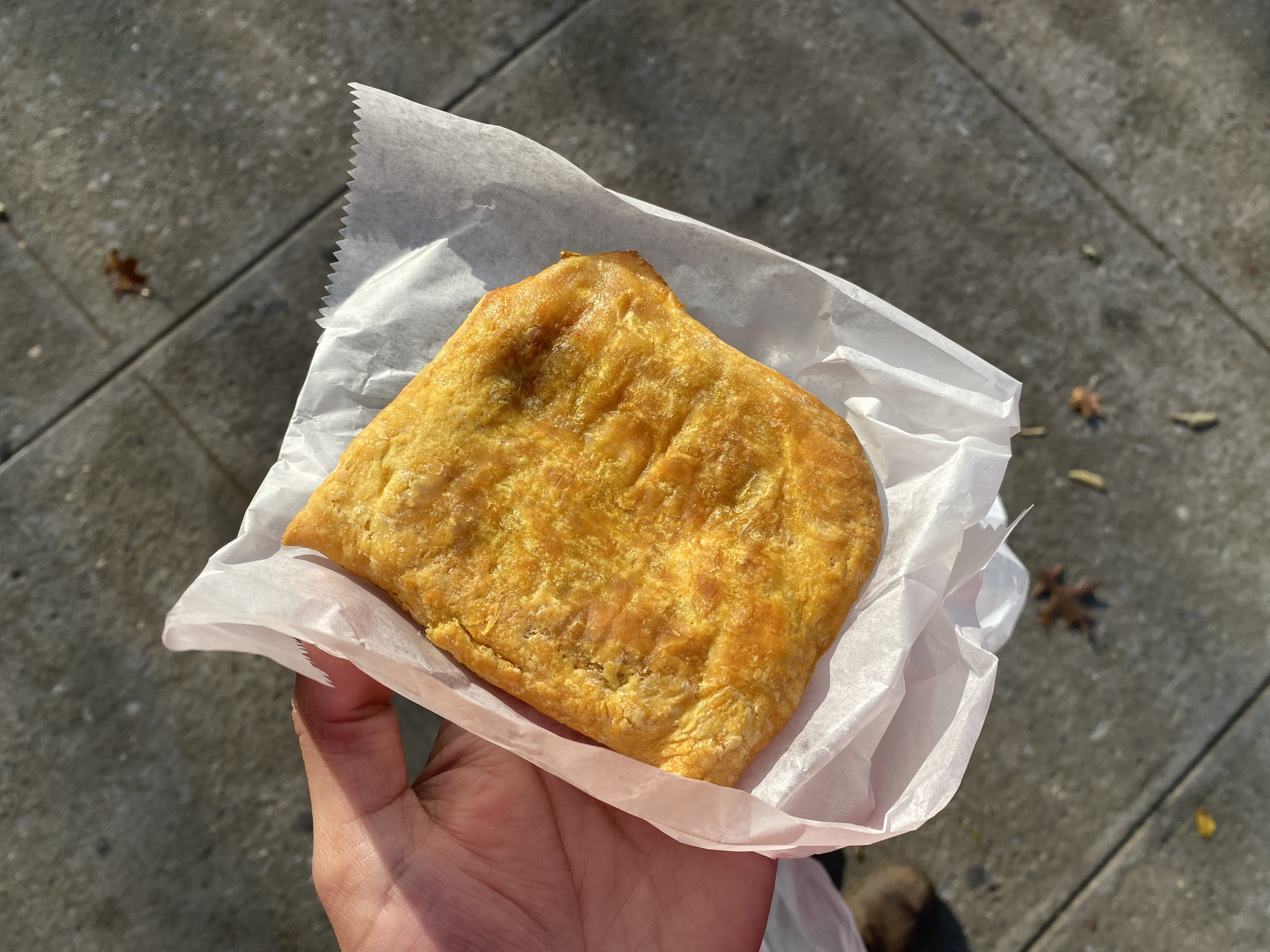 A hand holds a flakey curry chicken patty in a piece of parchment paper.