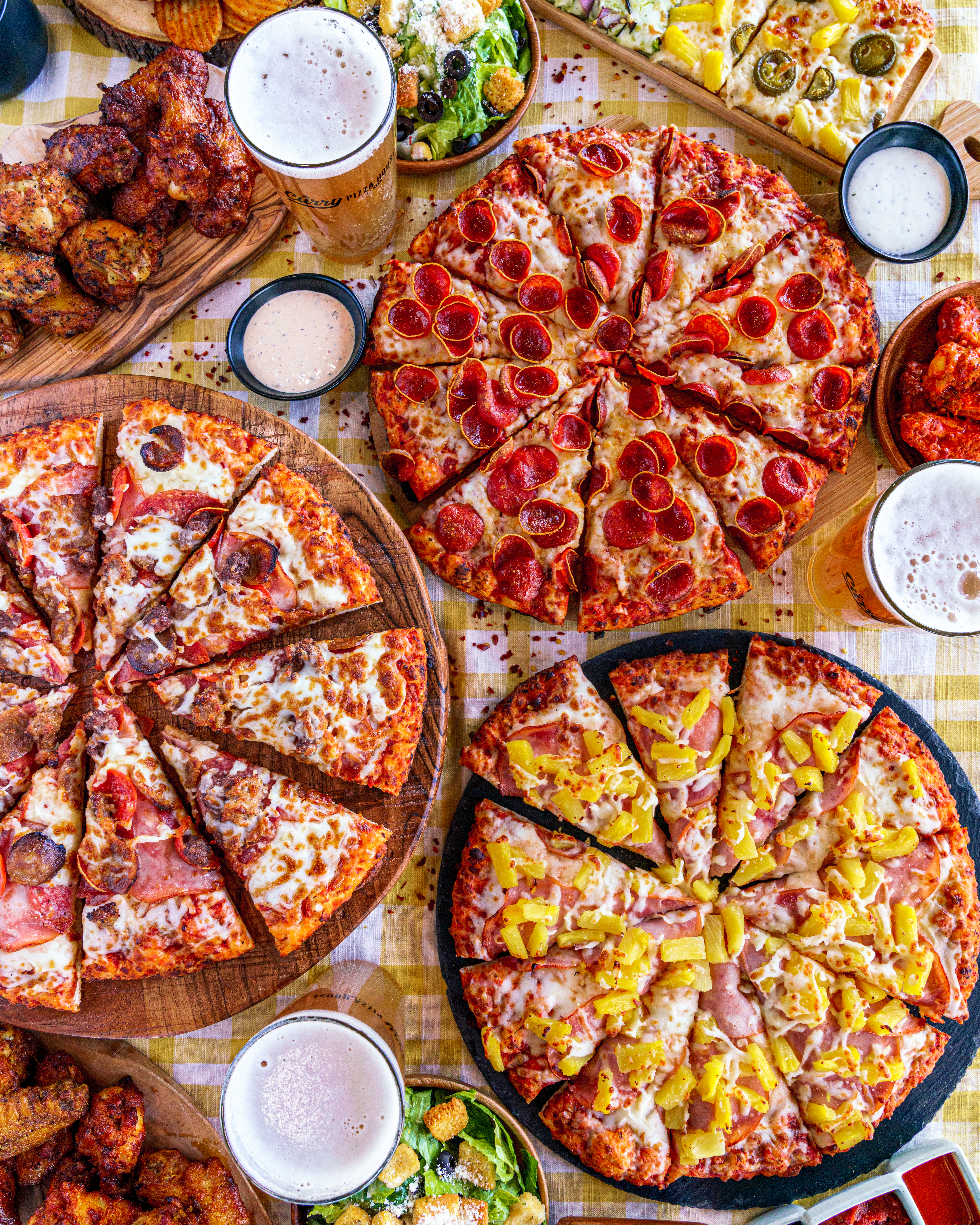 Three pizzas cut into slices - Hawaiian, Meat Lovers, and Pepperoni - on a table with beer