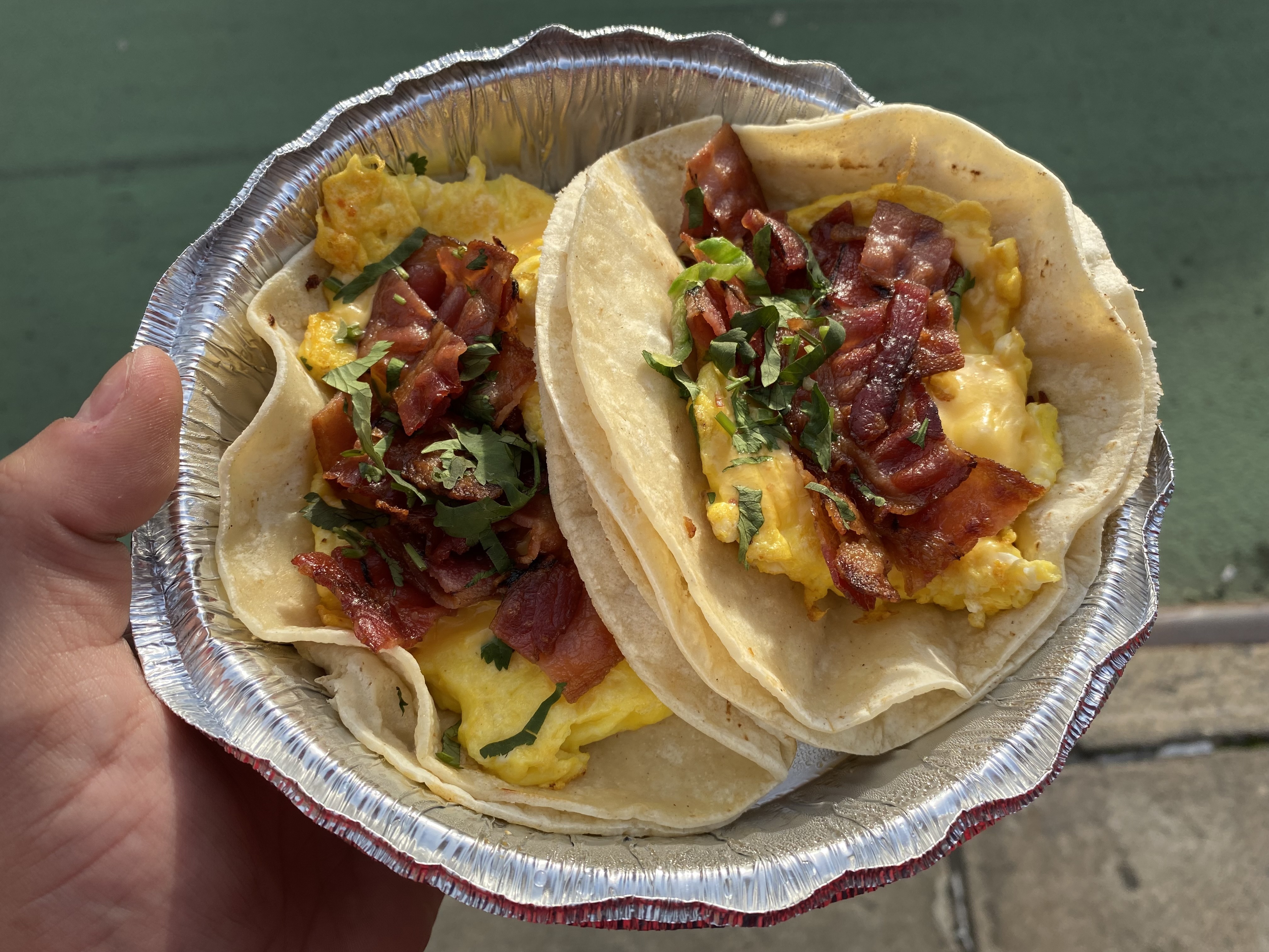 A hand holding a foil container with two tacos stuffed with bacon, egg, and cheese.
