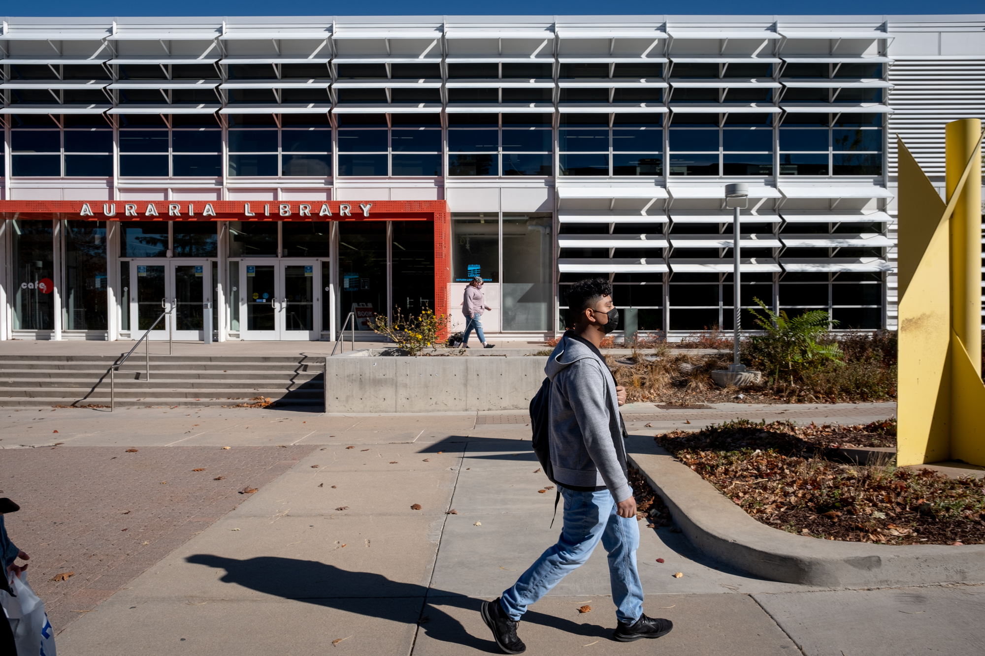 A young man, wearing a grey hoodie, jeans, and a black mask, walks on college campus past the Auraria Library.