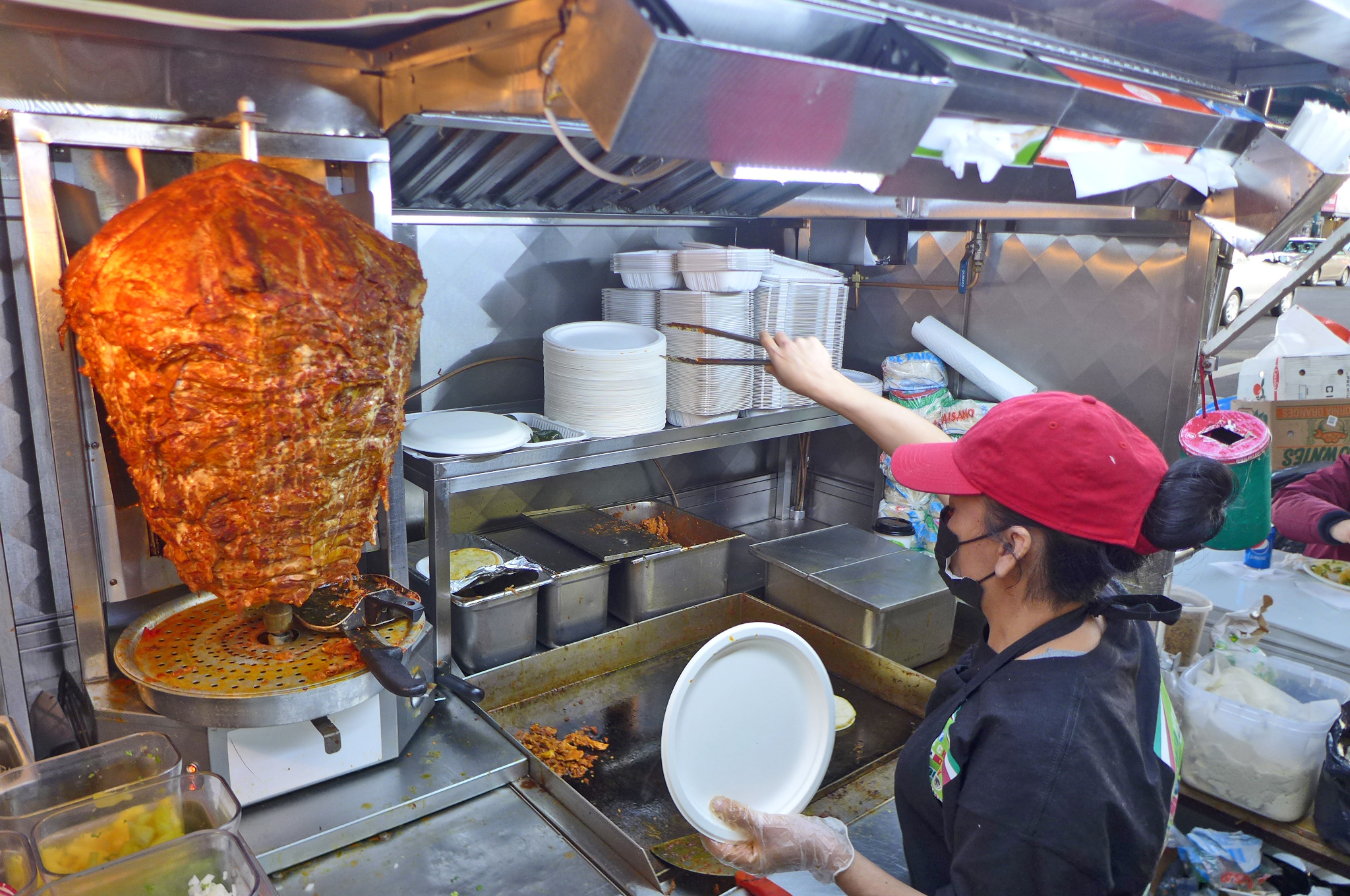 A big tapered cylinder of pork al pastor with a red-hatted lady in the foreground wielding tongs.