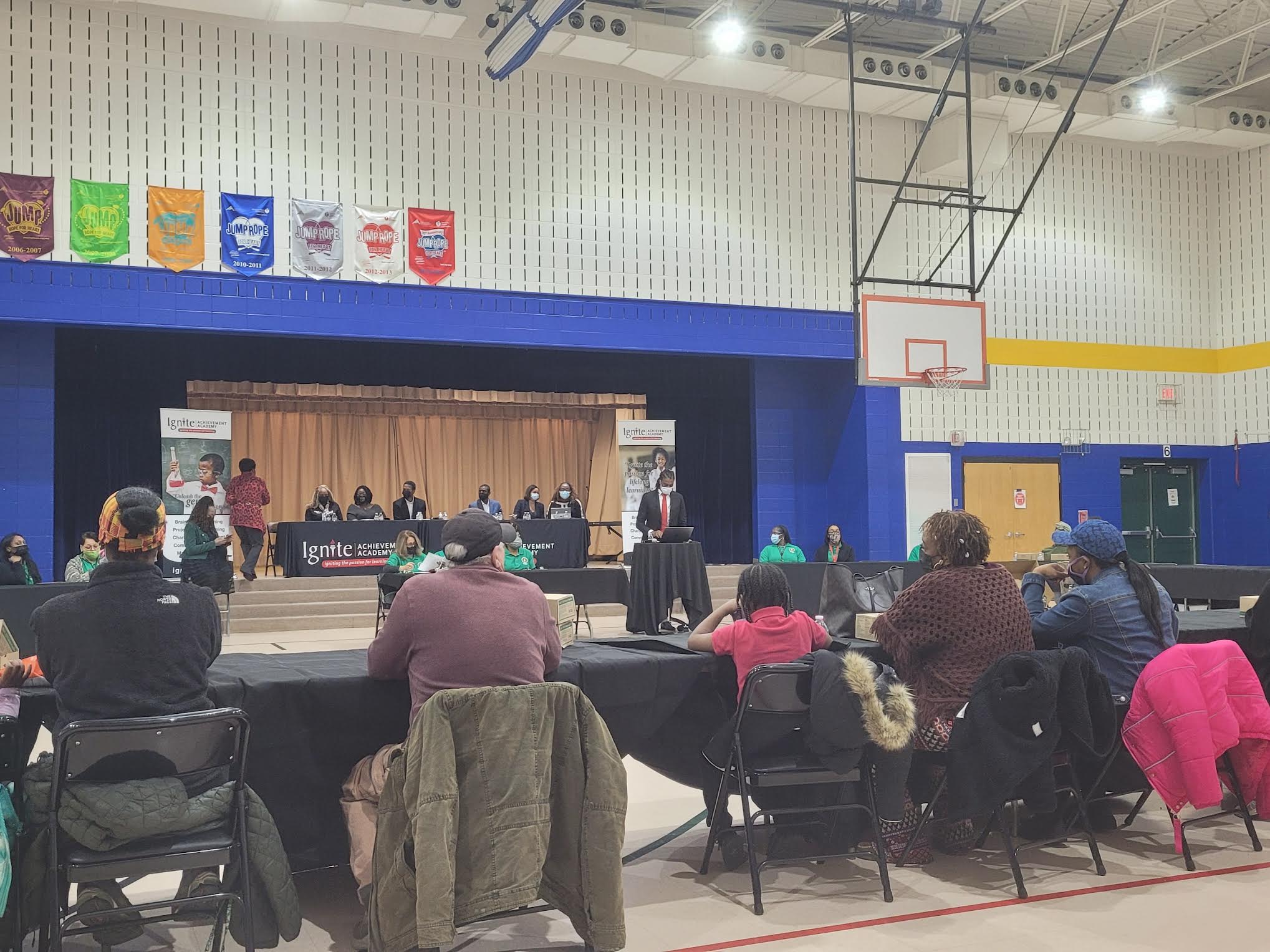 Four parents with their backs turned sit in a gym facing Ignite Achievement Academy co-founder Shy-Quon Ely as he gives a speech on the status of the school, with board members on a raised stage behind him.