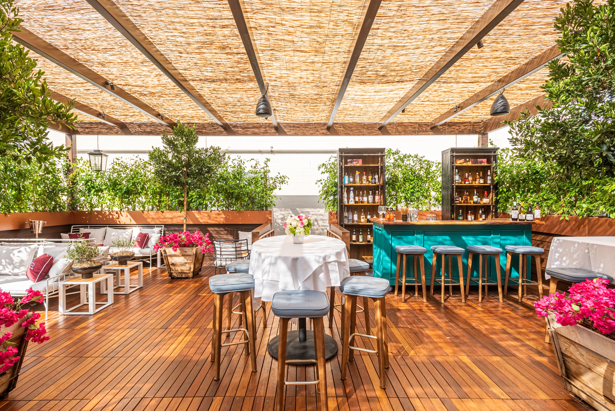 A daytime patio shaded from the sun with wood planks beneath, a teal bar, and lounge areas.