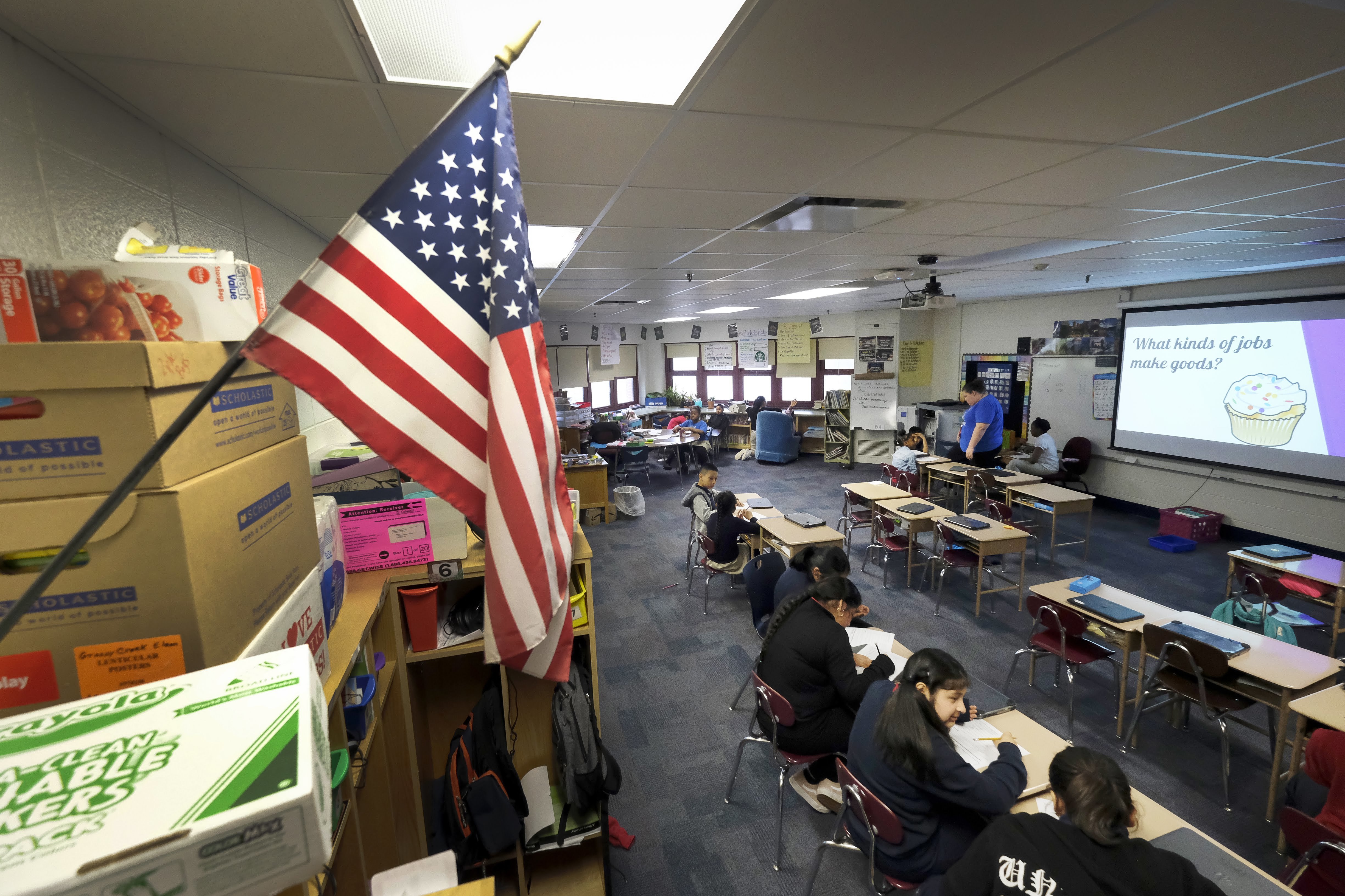 An American flag hangs on a wall in April 2019, in an elementary classroom with a few students sitting in a row