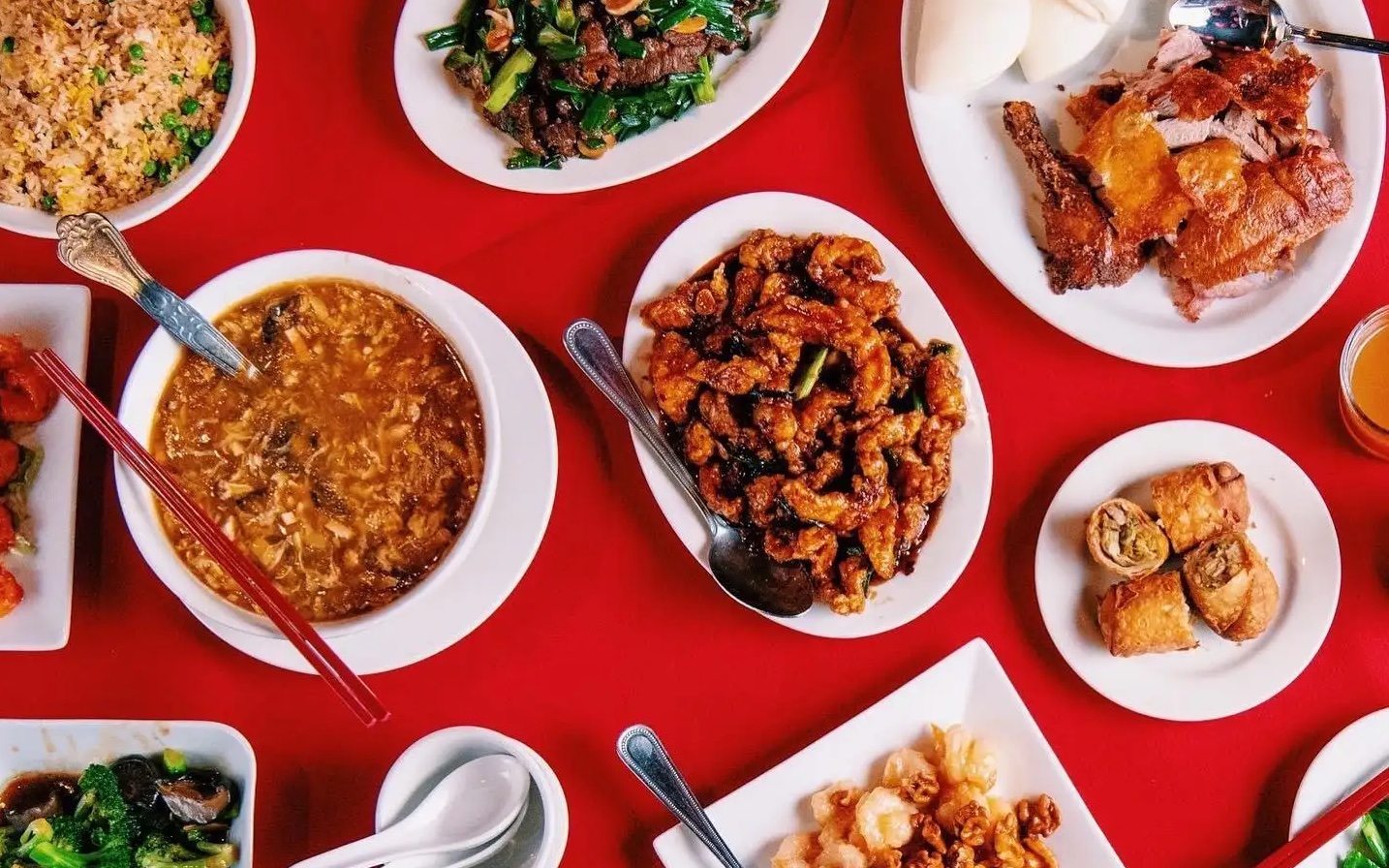 An overhead shot of white bowls and plates filled with Chinese food on a deep red table.