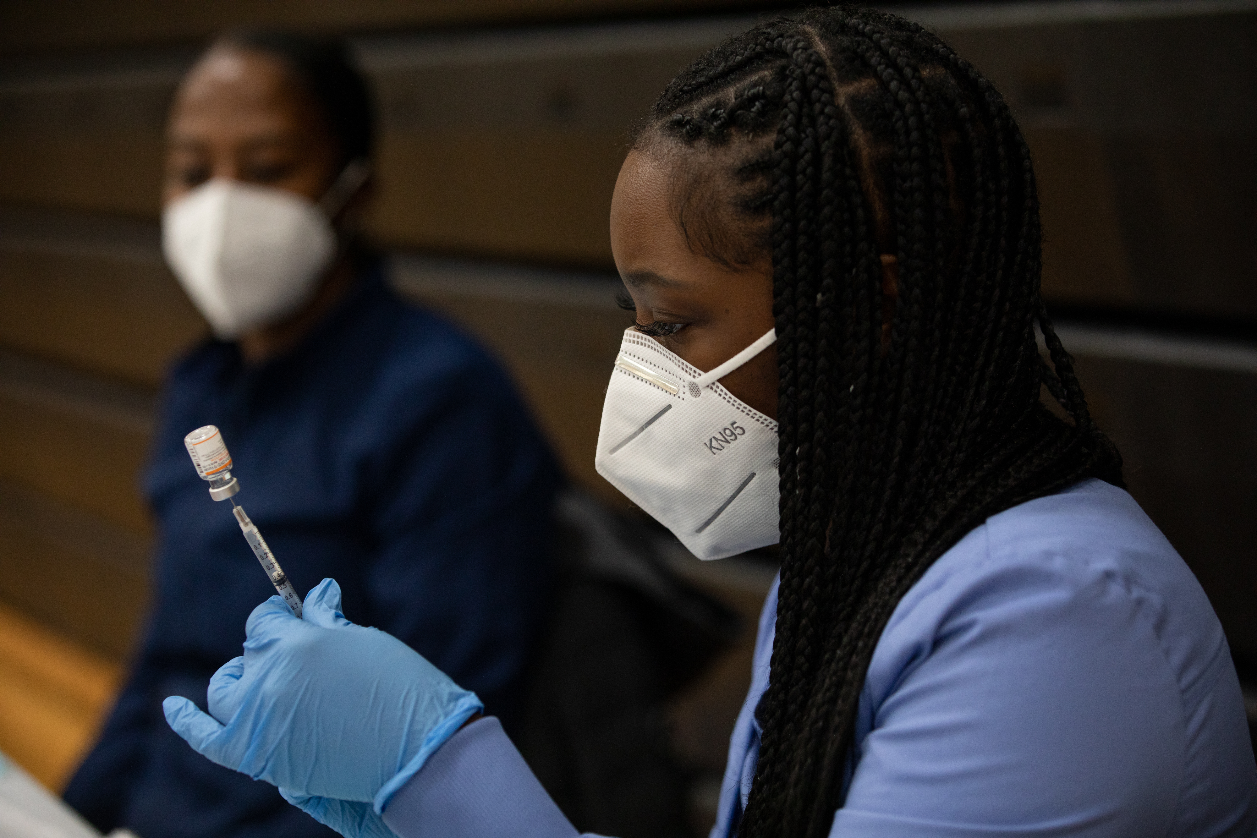 A woman wearing scrubs, medical gloves, and a KN95 mask draws vaccine from a vial using a syringe needle. Another person in the background watches.