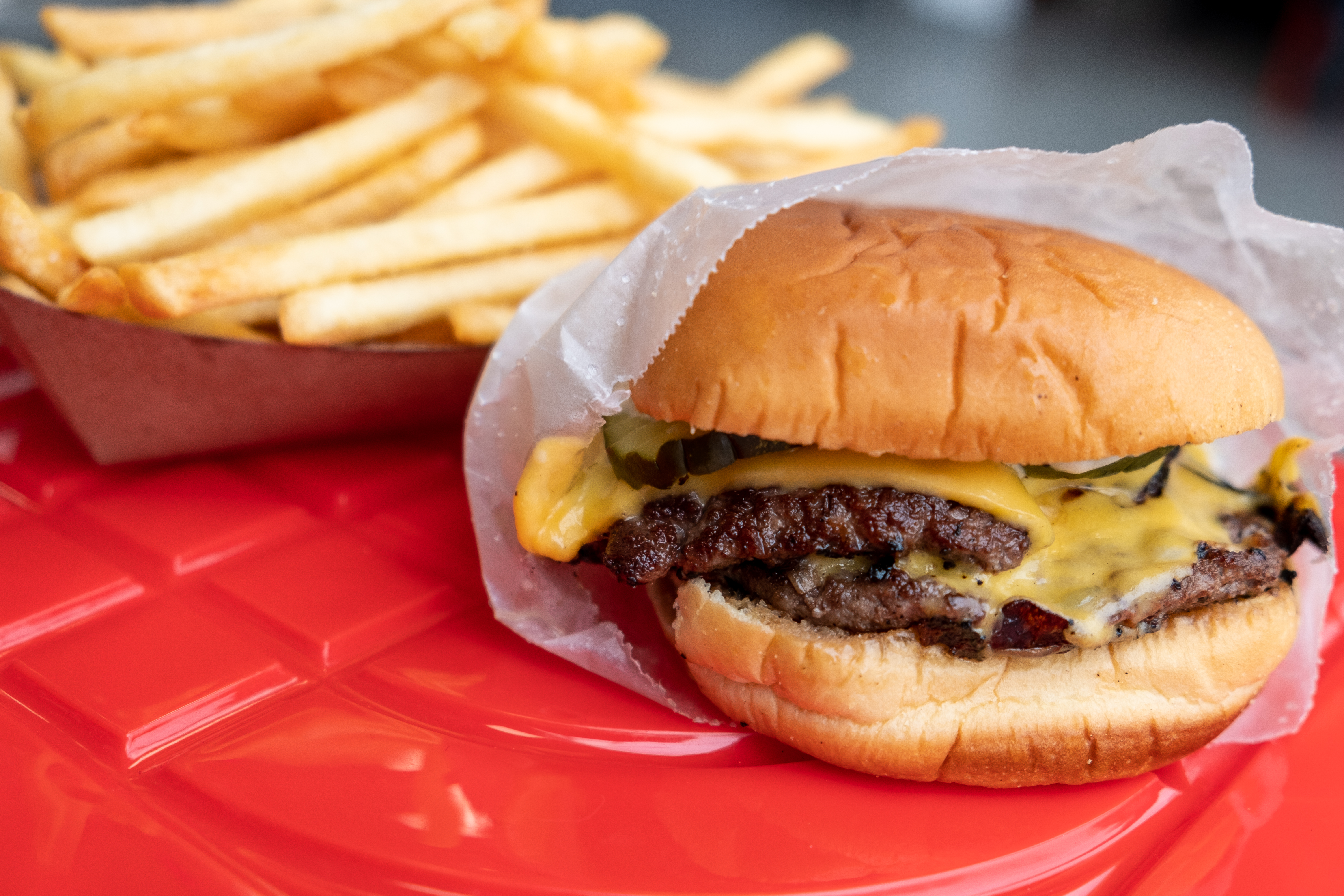 A double patty cheeseburger and fries sit on a red plastic stool in front of a restaurant. shown from up close.