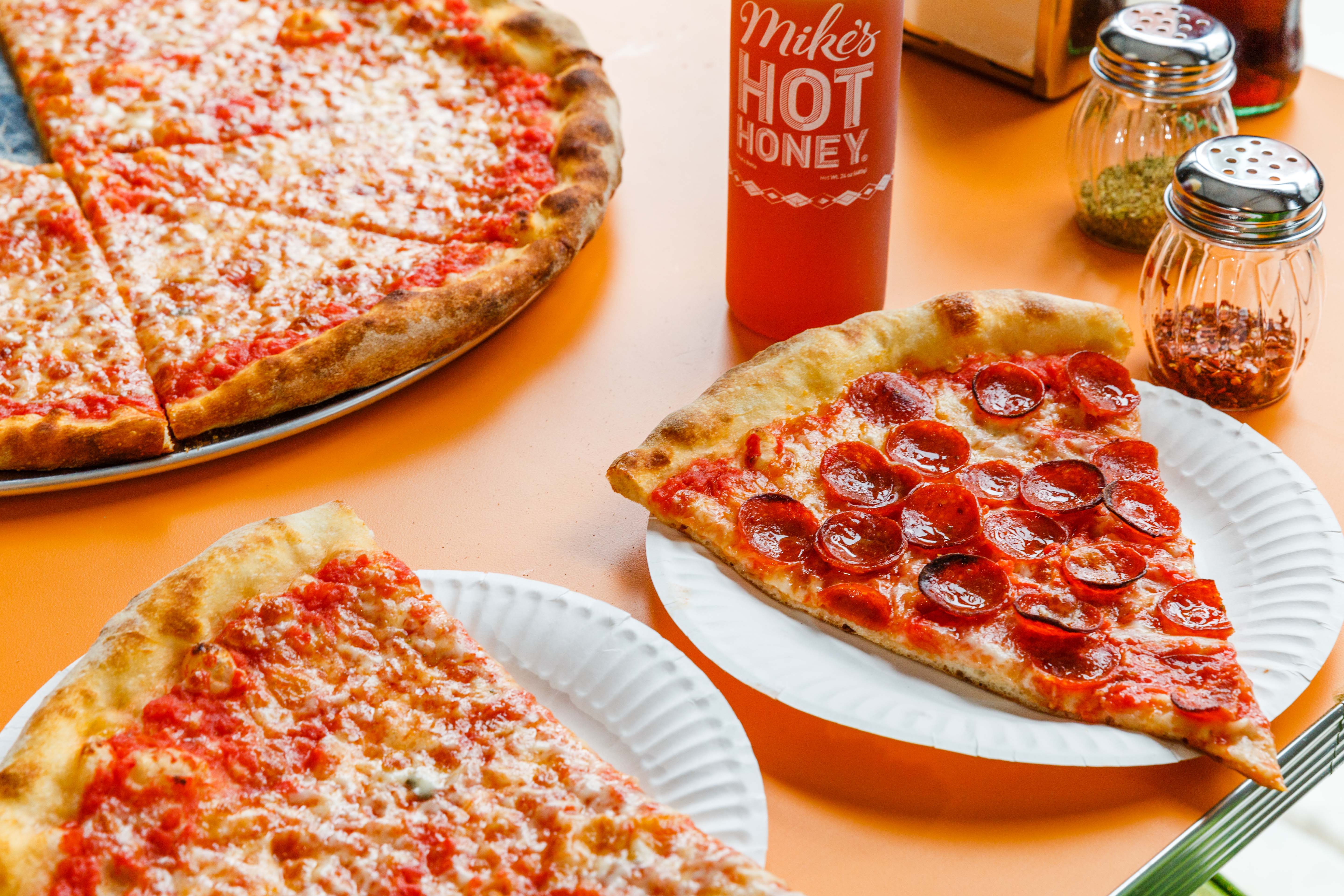Pepperoni and cheese slices sit on an orange countertop at Paulie Gee’s slice shop, adjacent hot honey sauce and chile flakes