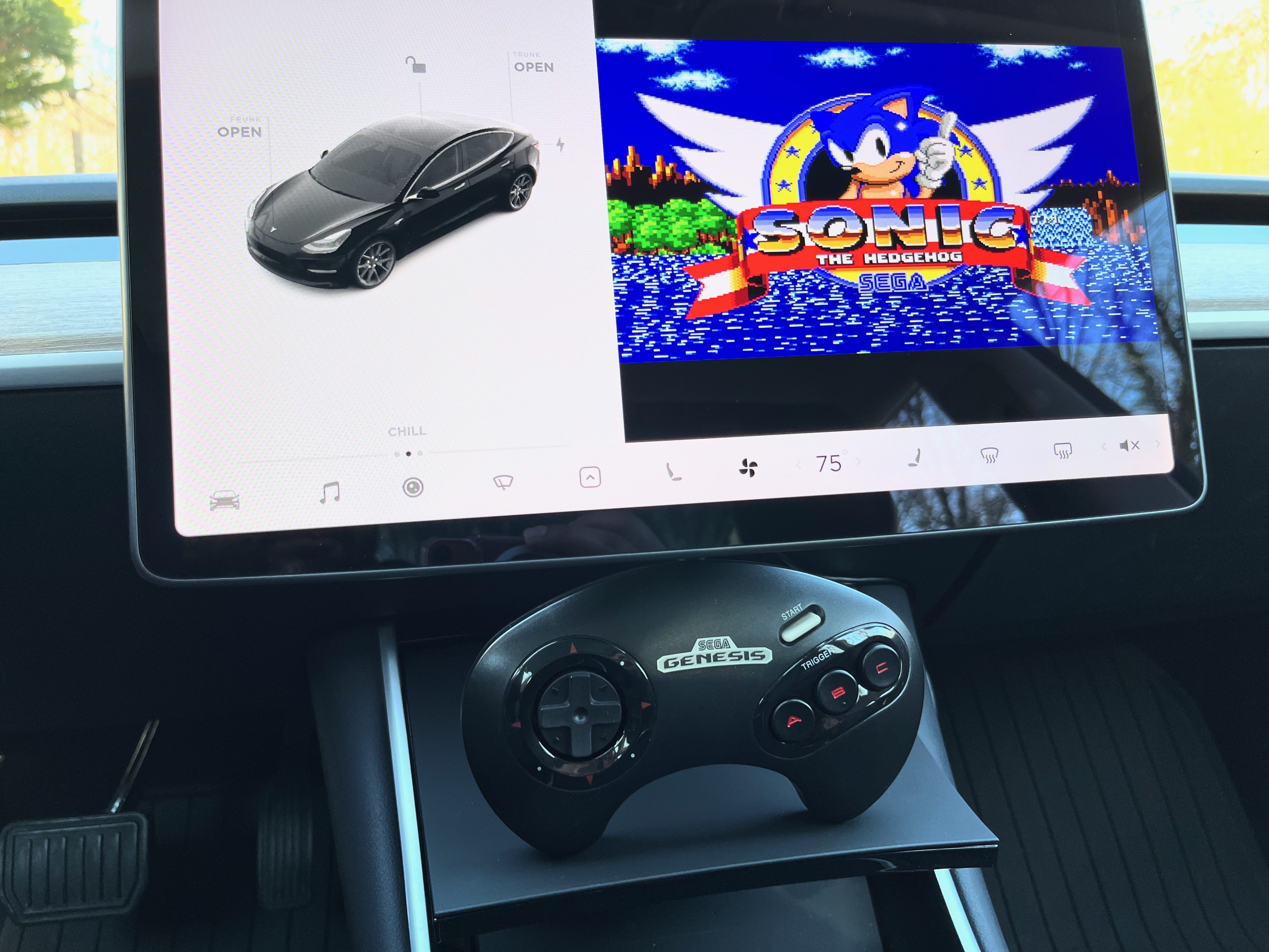The Tesla Model 3 interior is displayed with a Sega Genesis game controller sitting under the infotainment screen with a window on the screen showing the game Sonic 1.