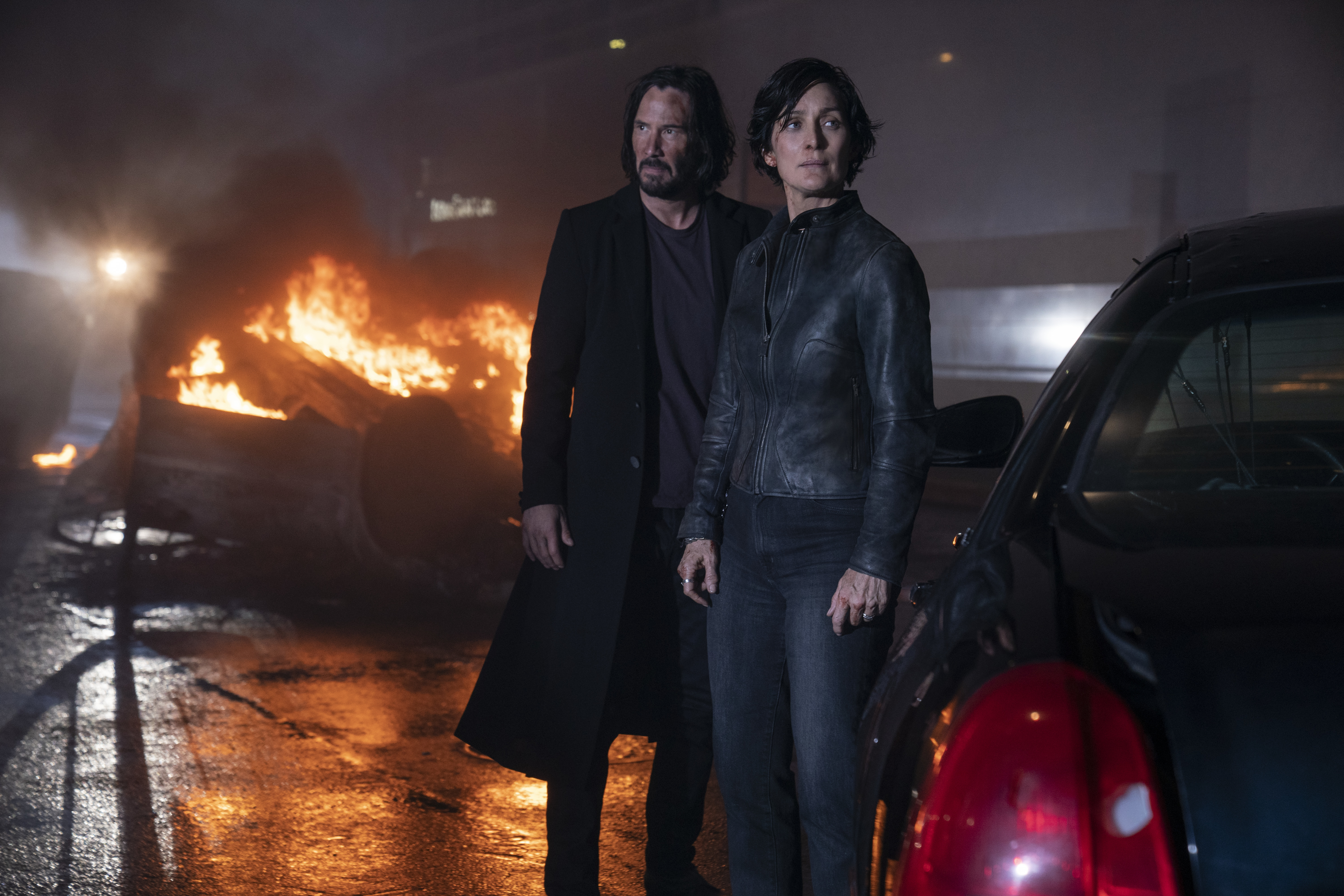 Neo and Trinity stand in front of burning wreckage in The Matrix Resurrections.