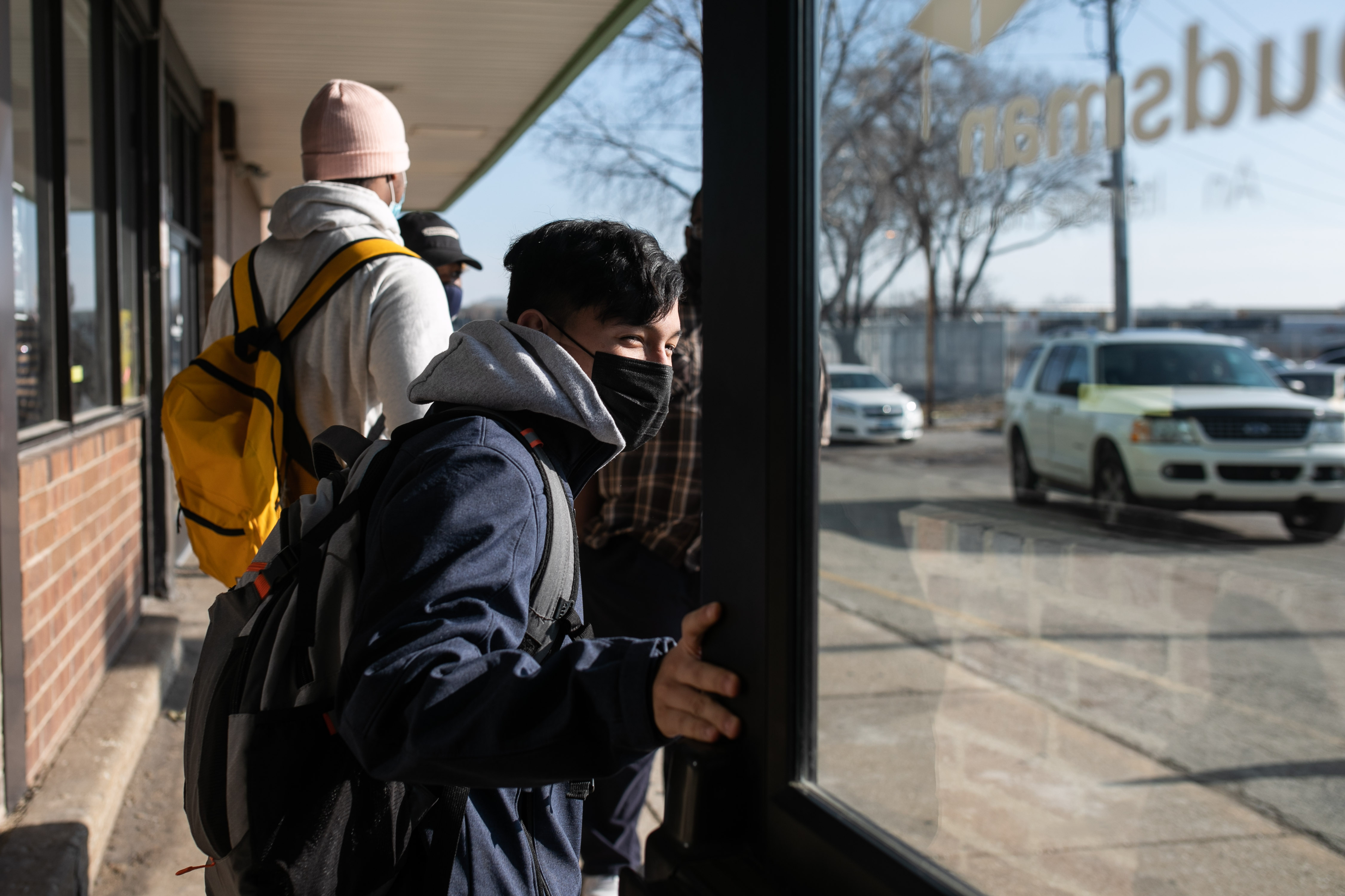 A student, wearing a black mask, blue jacket, and grey hoodie, holds a door open as he exits school. Other students are on the sidewalk directly behind him.
