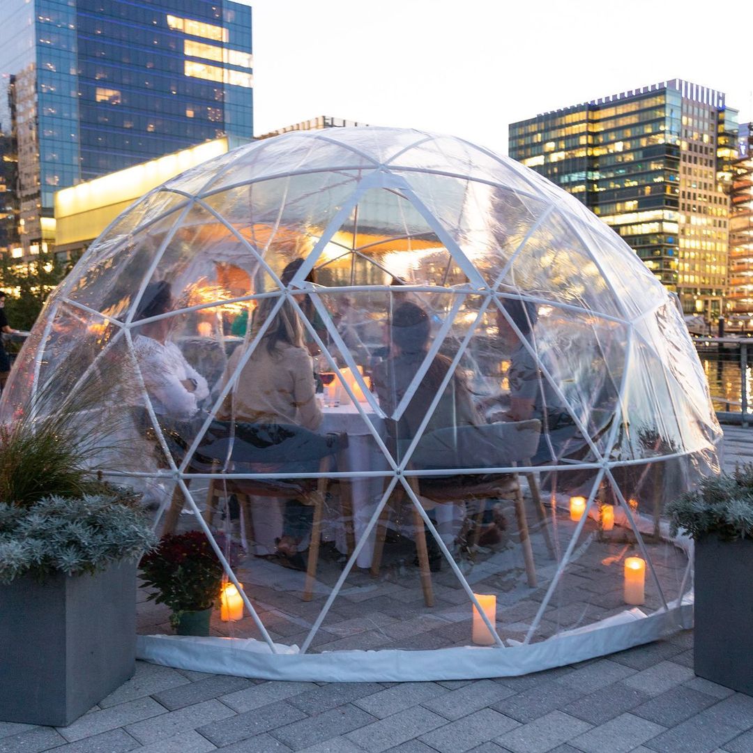 Outdoors at dusk, a see-through igloo is full of people dining. City buildings are visible in the background.