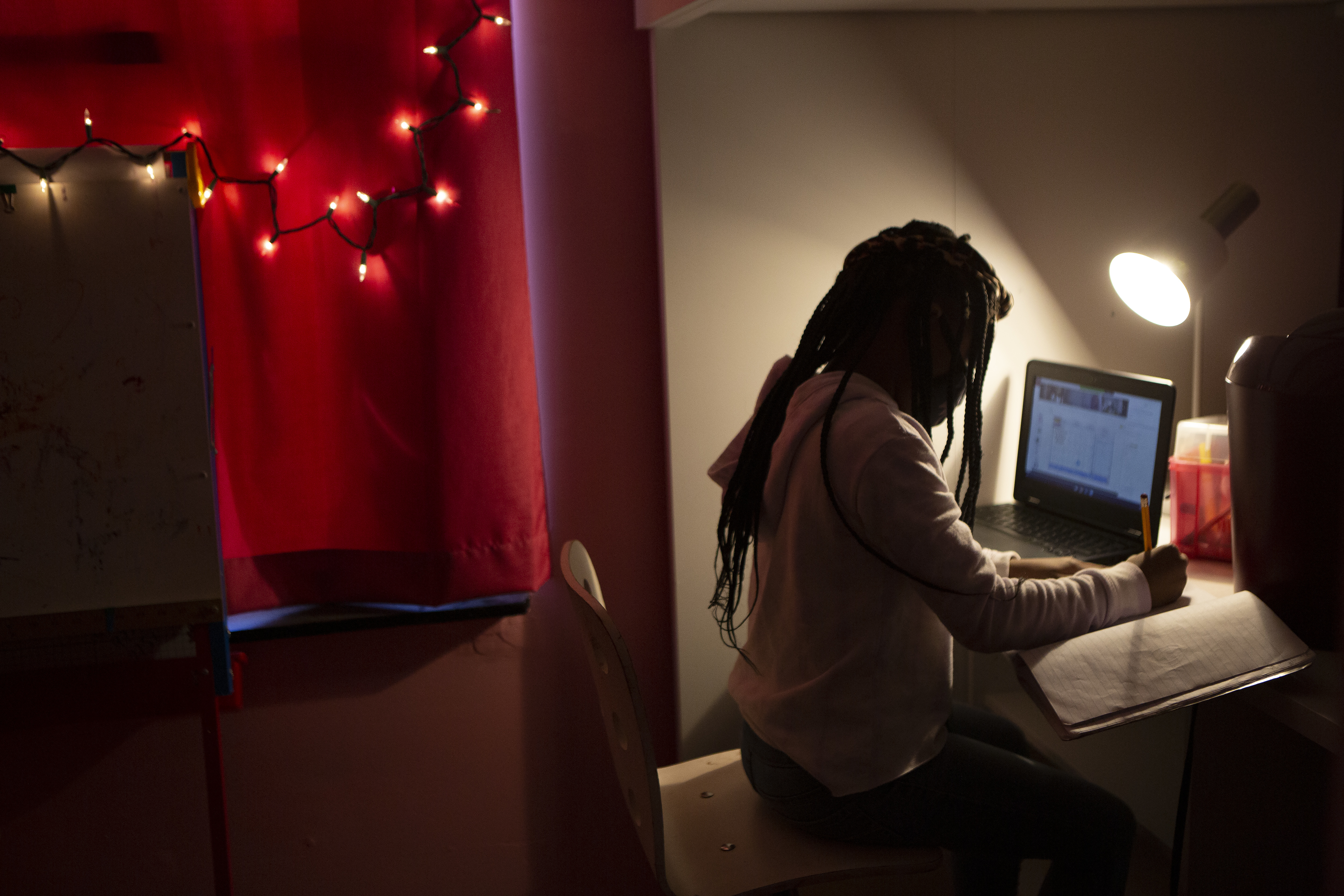 A young girl in a darkened room works in the light of a desk lamp. She is writing in a notebook. A laptop computer is open on her left. Behind her a string of Christmas lights hang over a red curtain.