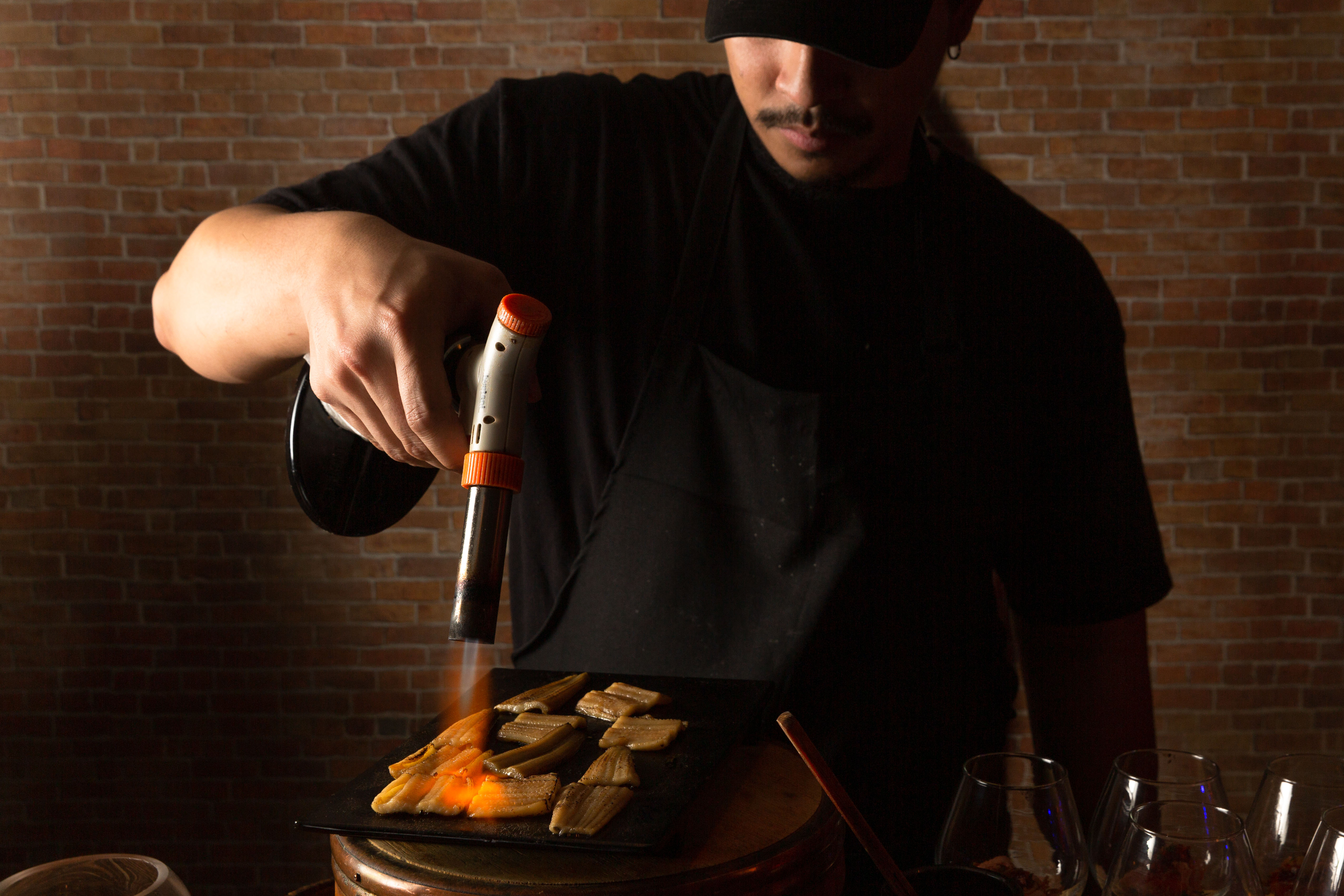 A chef in a black shirt holds a blowtorch over a pile of brown unagi